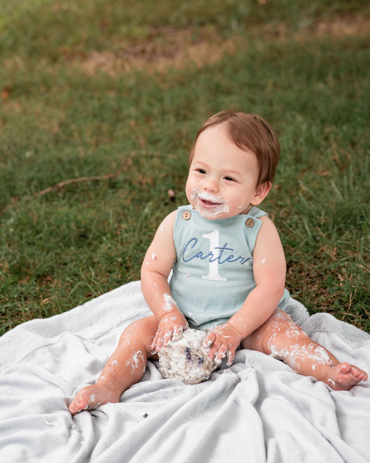 Baby Carter smiles with cake on his face during a first birthday cake smash photoshoot outdoors on a blanket.