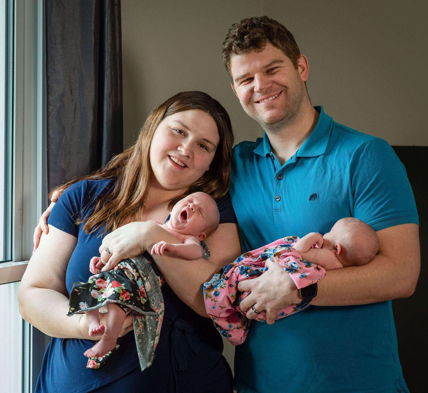 Happy couple smiling and holding two newborn babies wrapped in colorful blankets near a bright window.