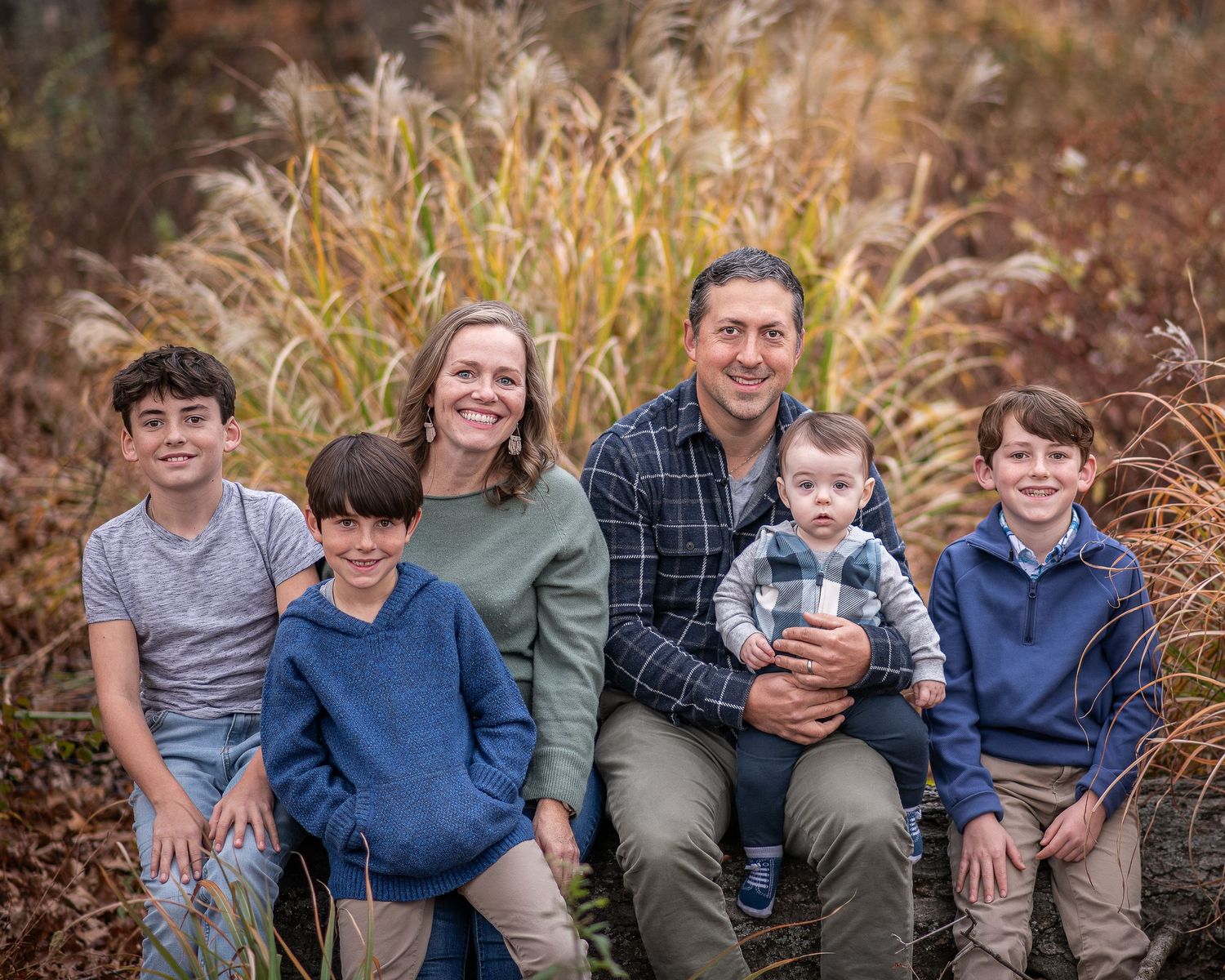 Family of six posing outdoors in autumn, parents with four boys sitting together among golden ornamental grasses.