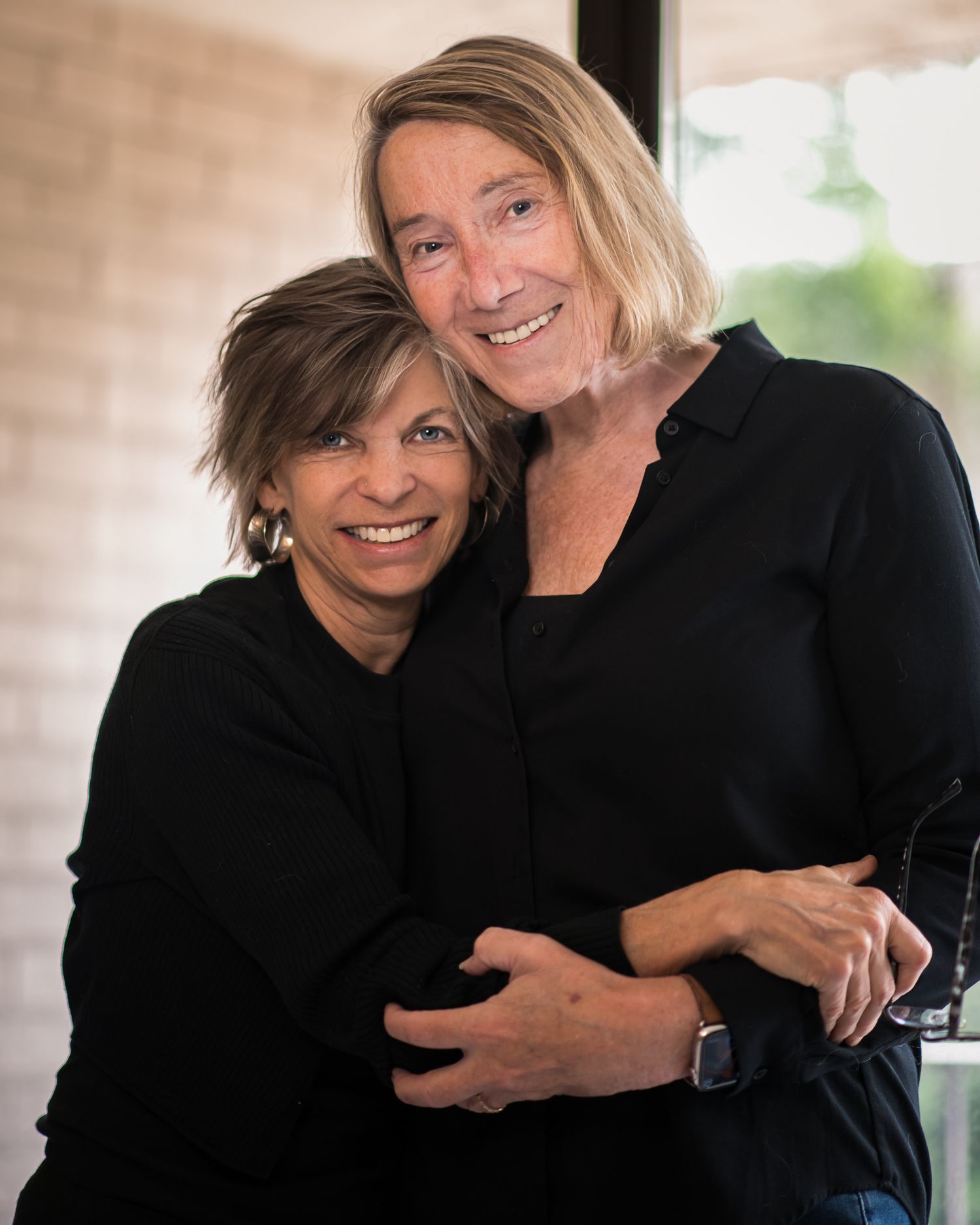 Two smiling women in black tops embrace warmly in a bright indoor setting, sharing a joyful moment together.