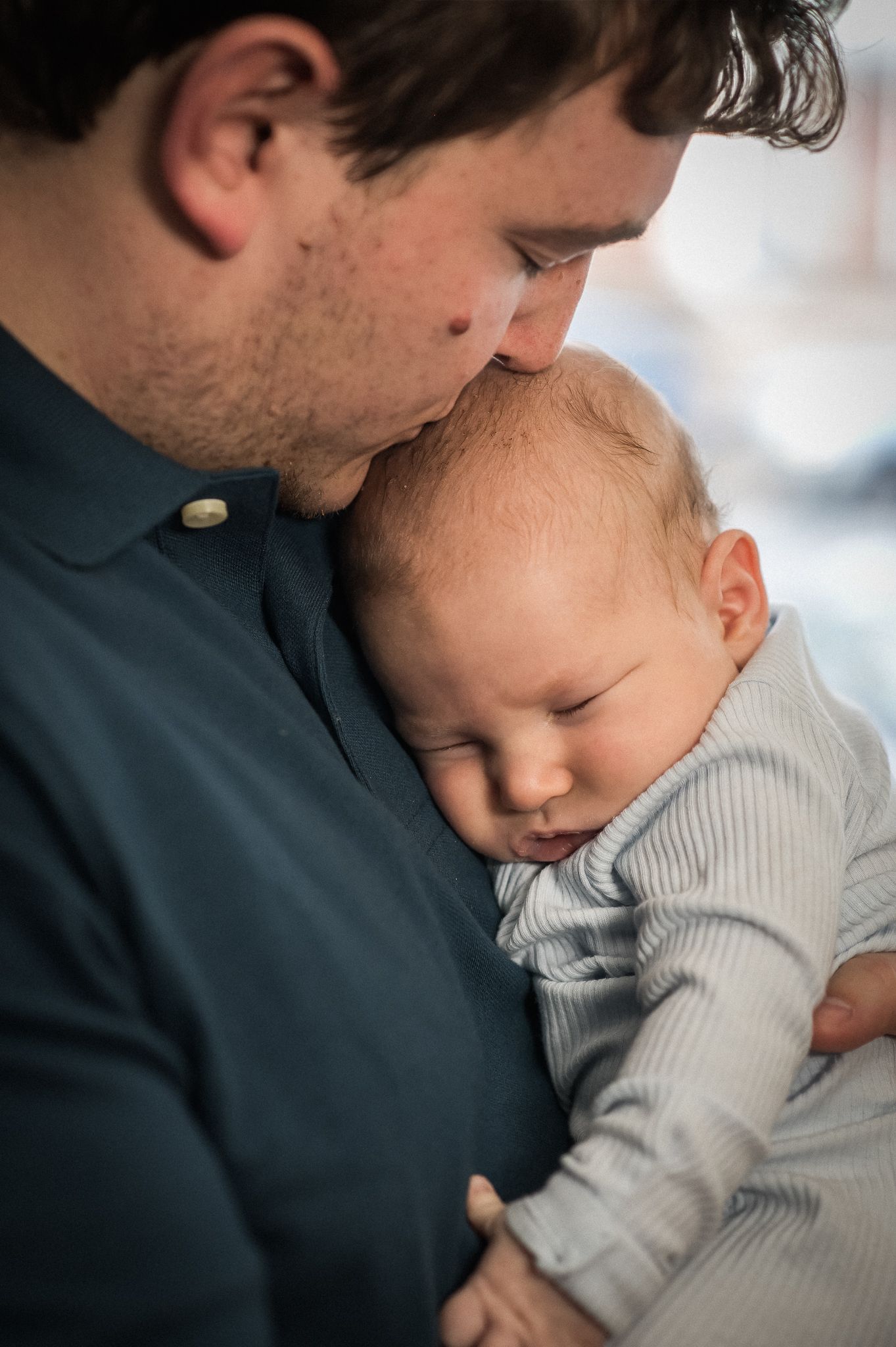 A tender moment shows a sleeping baby in a gray onesie resting peacefully against someone in a dark shirt.