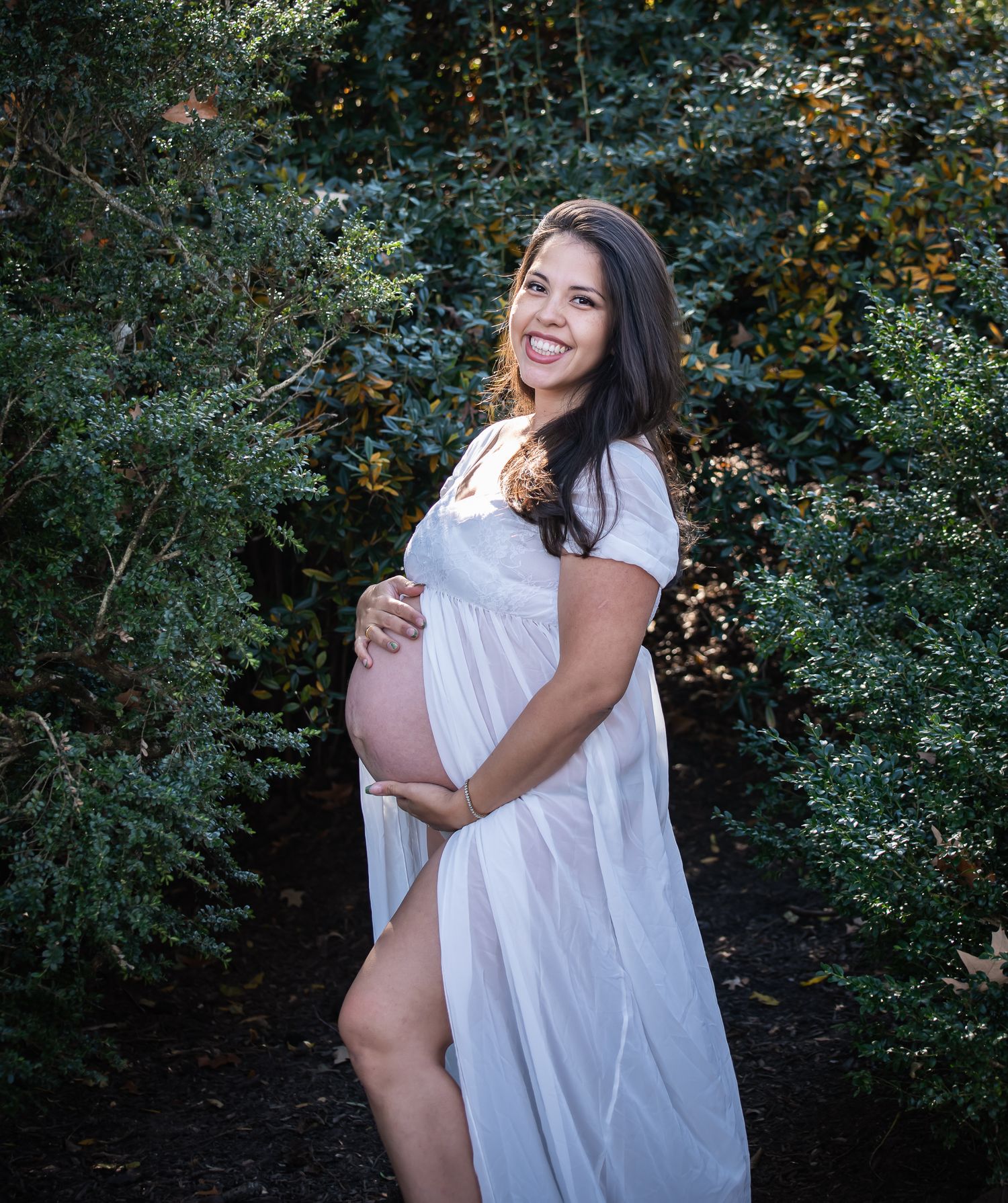 A maternity photoshoot in a lush garden setting with soft natural lighting and white flowing fabric.