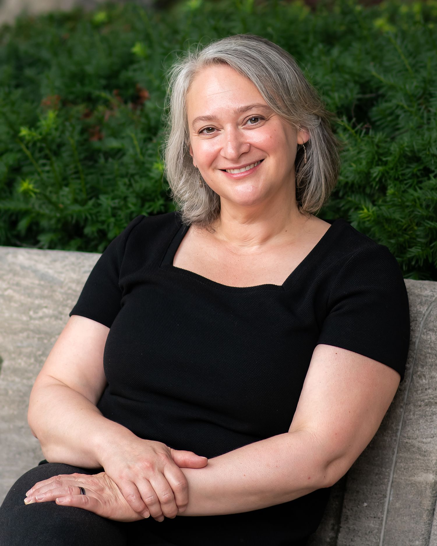 A smiling woman with gray hair wearing a black top sits outdoors in front of lush green foliage.