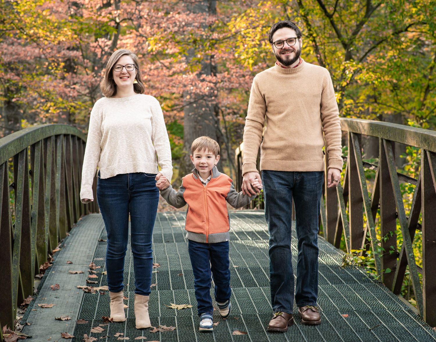 A family walks together holding hands on a wooden bridge surrounded by colorful autumn foliage.