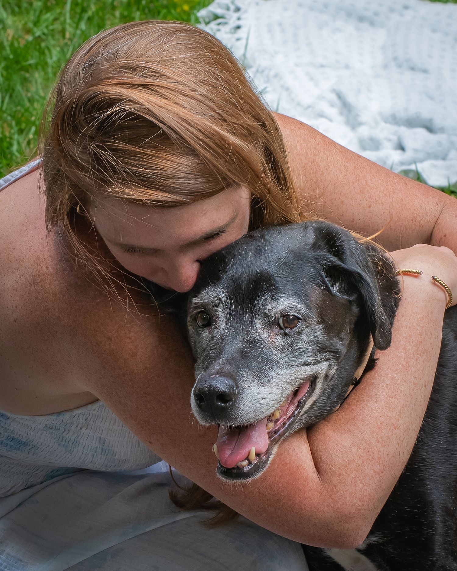 A woman with strawberry blonde hair hugs and kisses the head of a senior black dog with a gray muzzle while sitting outdoors on the grass.