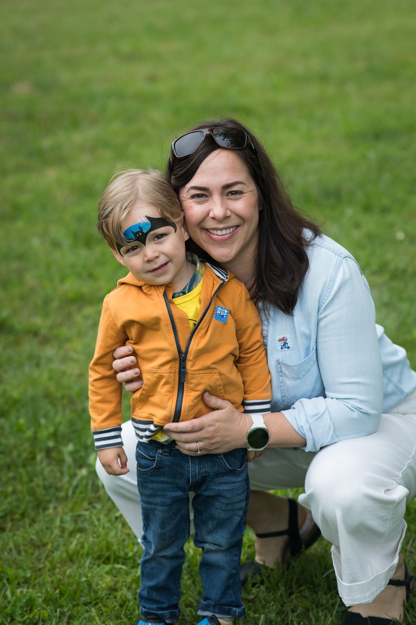 A smiling woman crouches beside a young boy with face paint on a green lawn, both posing happily together outdoors.