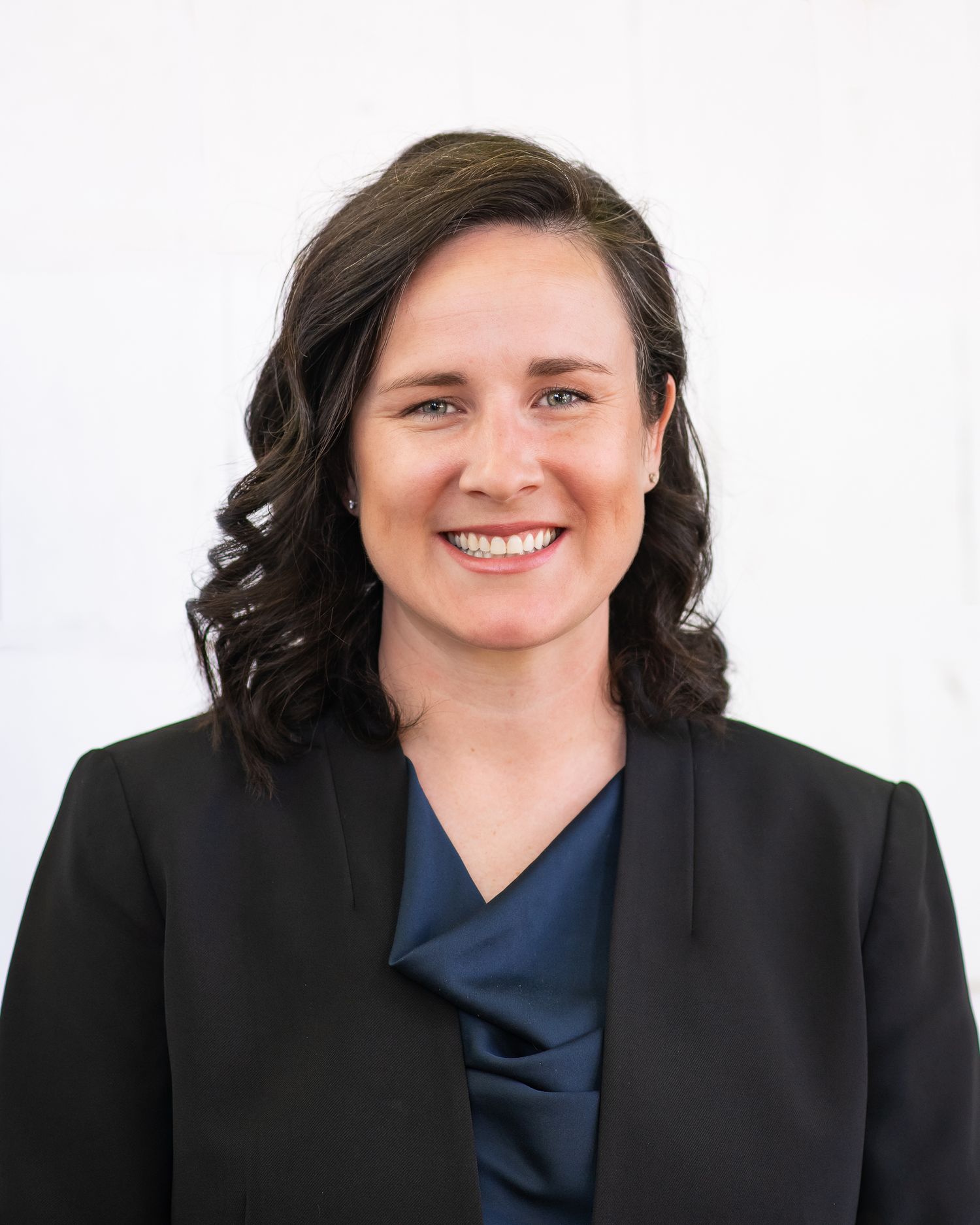 Professional headshot of a smiling woman with dark curly hair wearing a black blazer and teal blouse.
