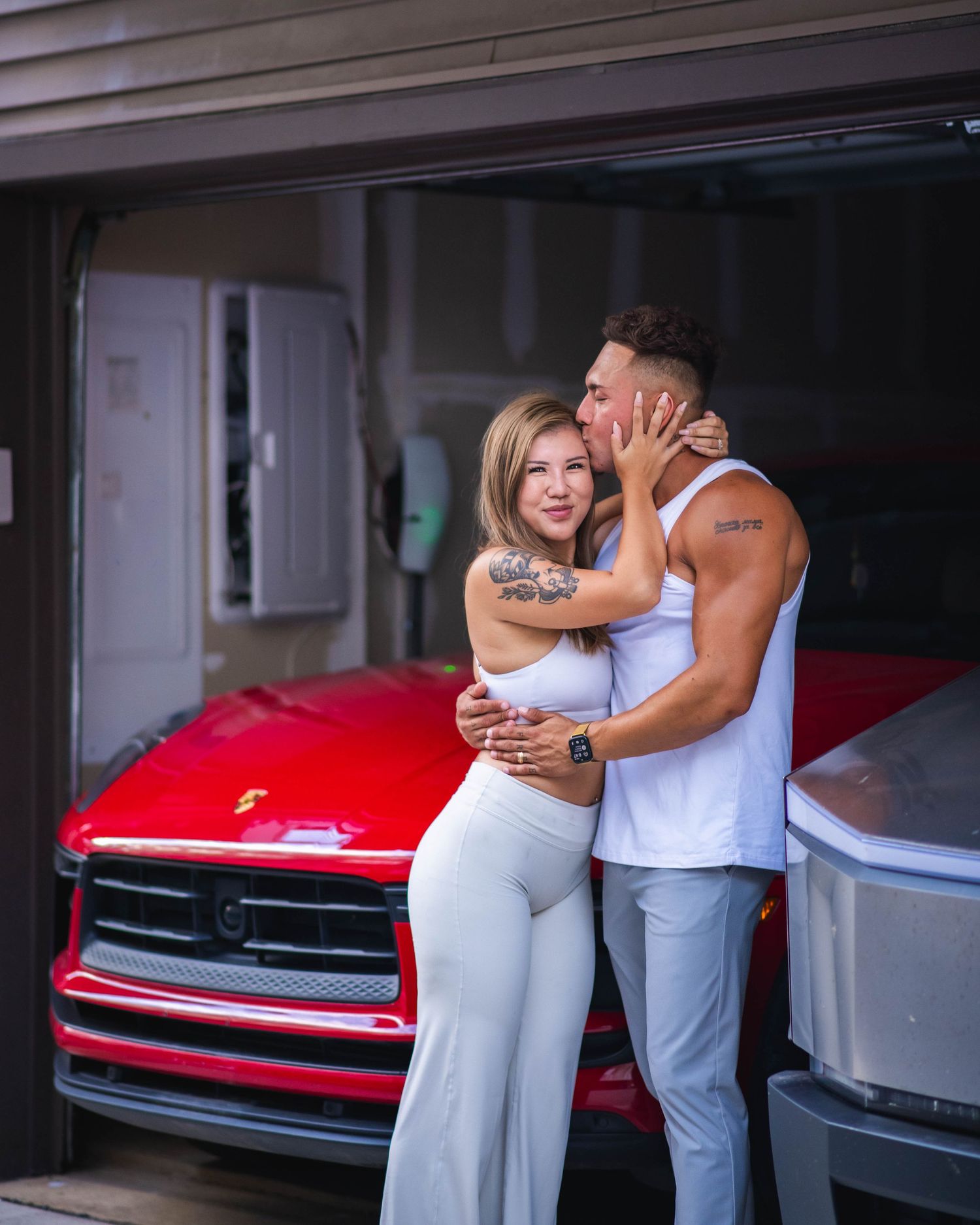 A couple embraces and kisses in front of a red truck in a garage, both wearing white outfits with visible tattoos.
