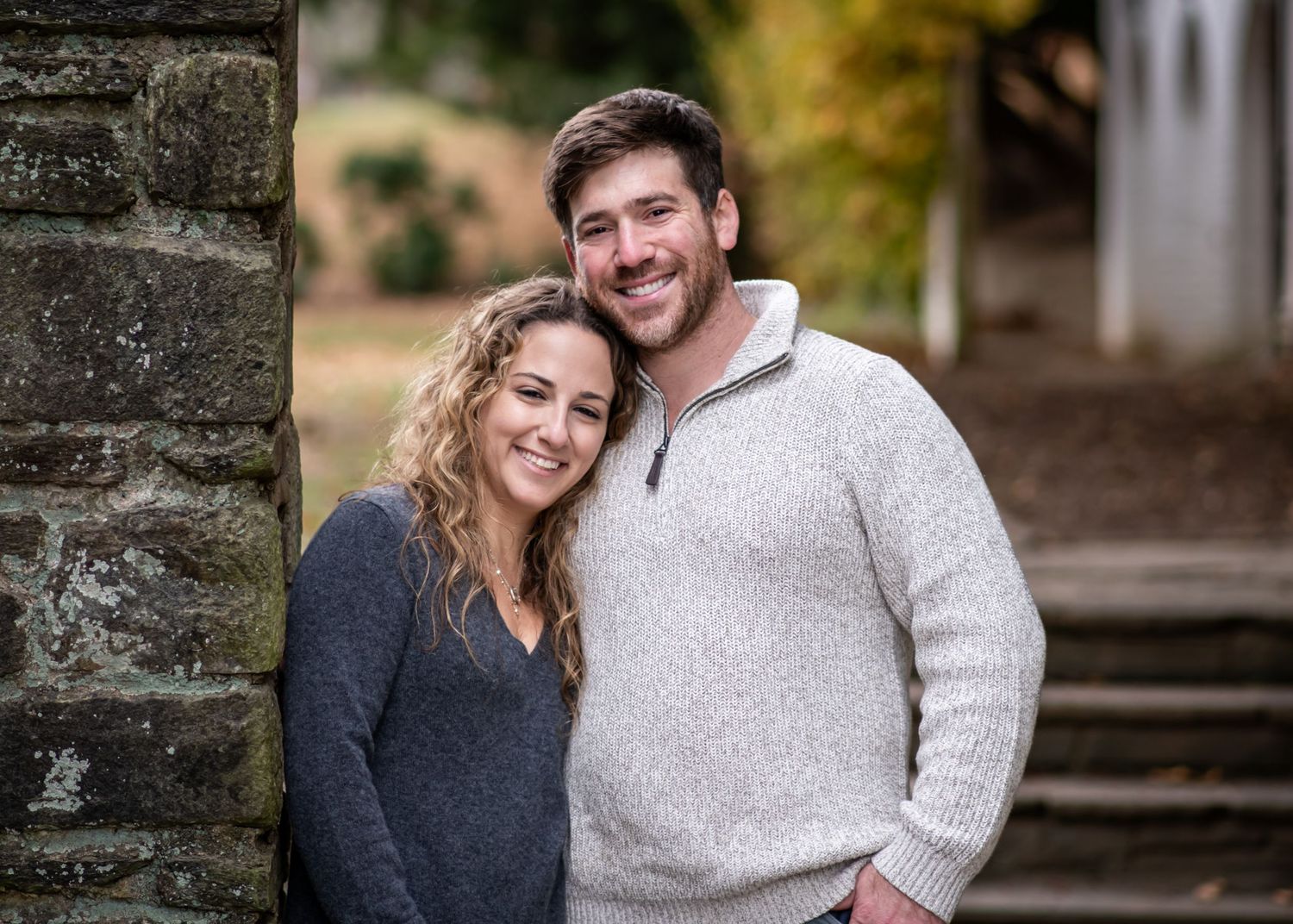 A couple wearing gray and navy sweaters embrace and smile while standing outdoors near a stone wall.