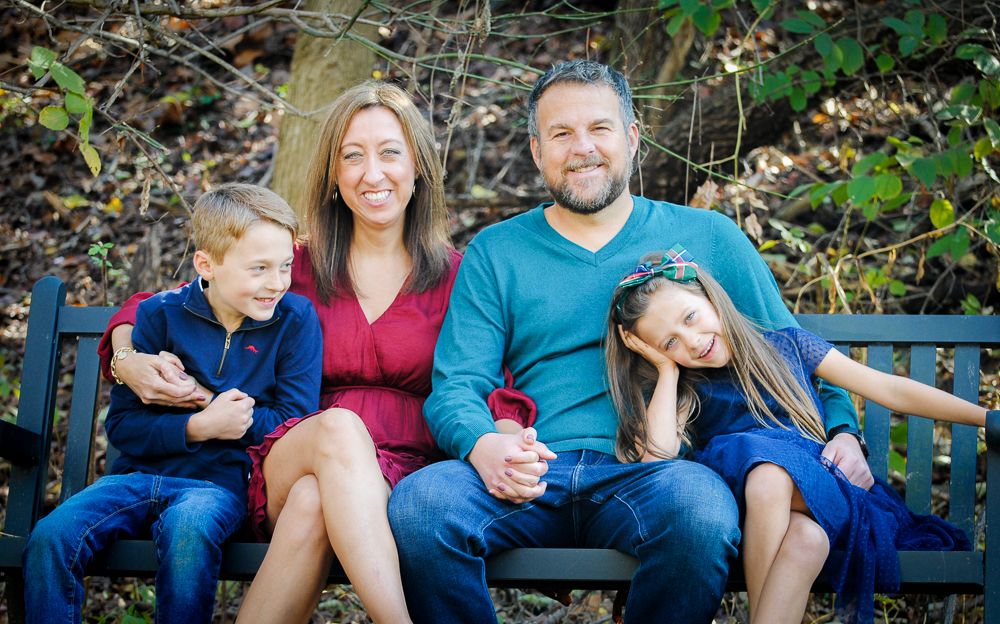 A family of four sits together on a wooden bench outdoors, smiling and enjoying a casual moment in nature.