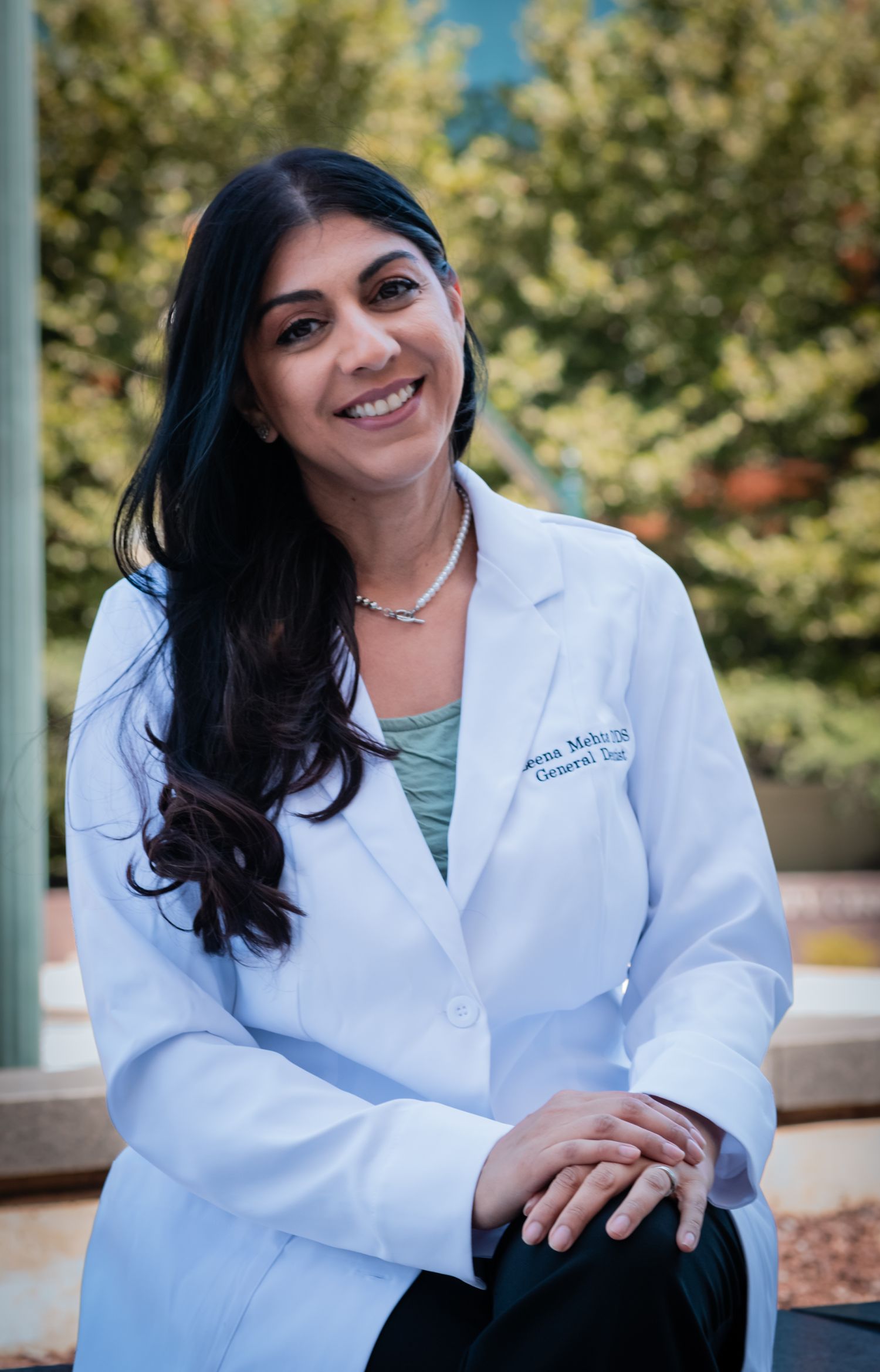 Healthcare professional in white lab coat sitting outdoors smiling against natural foliage background.