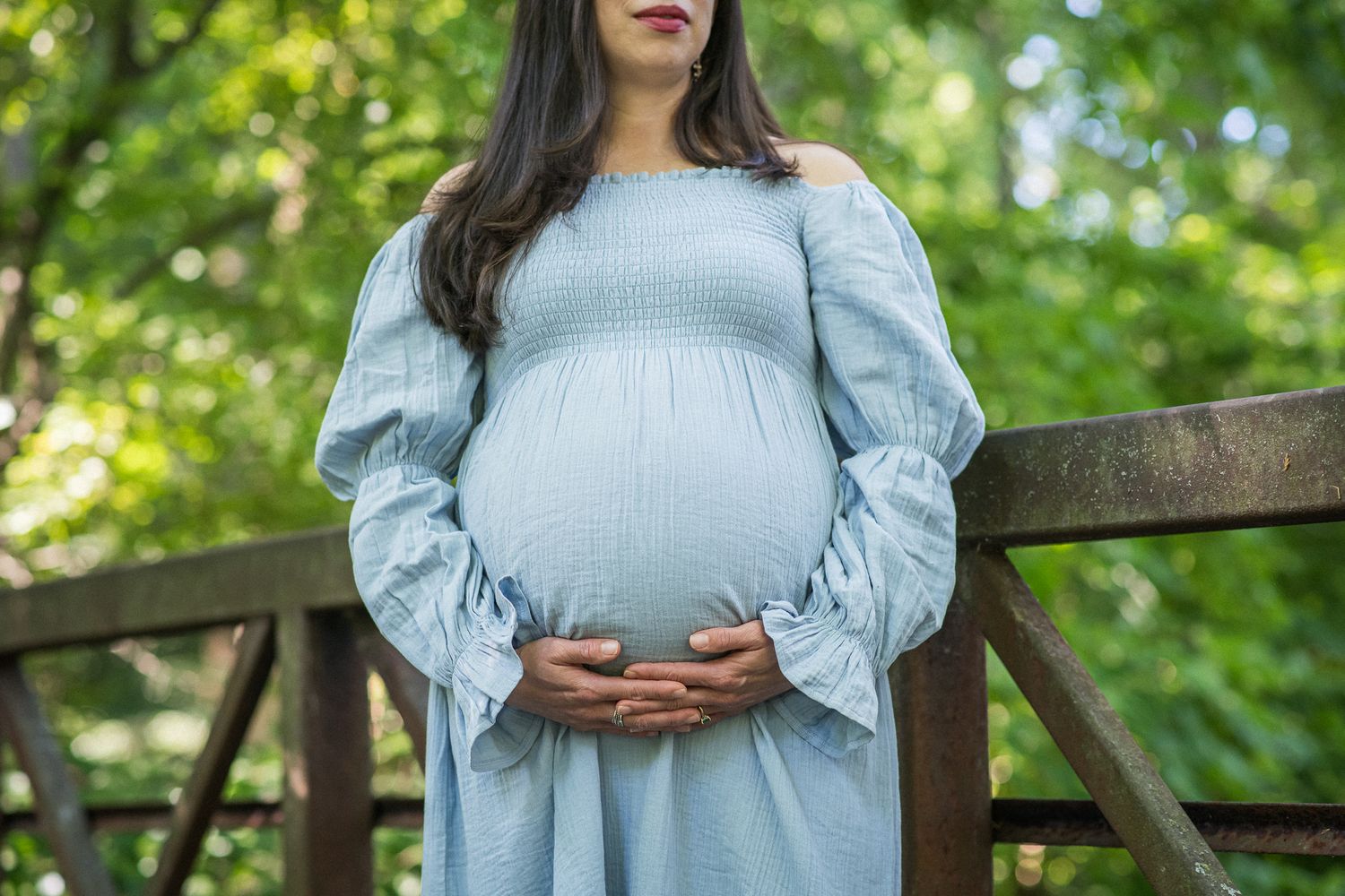 Pregnant woman in light blue dress cradling baby bump outdoors near wooden fence surrounded by lush green trees.