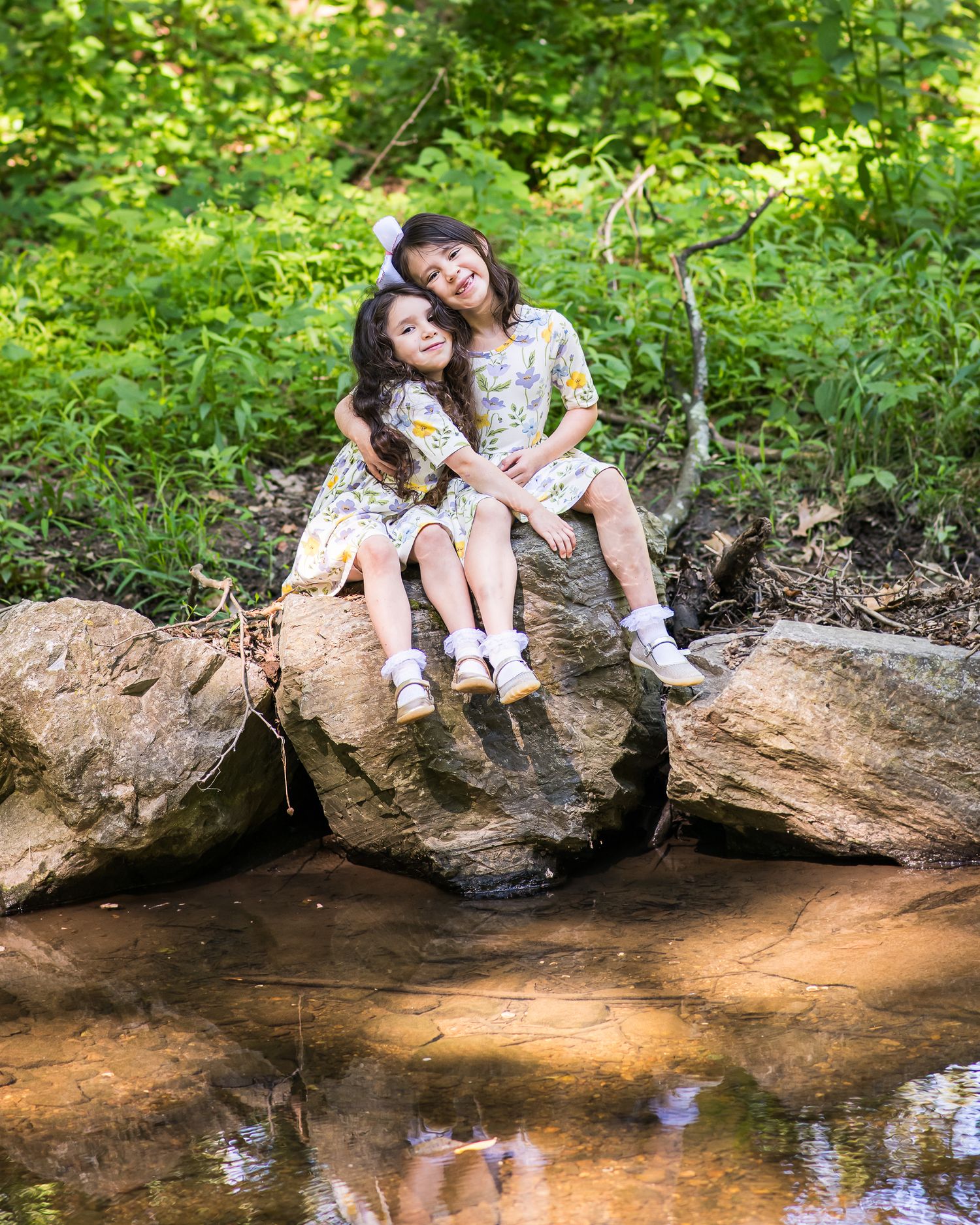 Two girls in floral dresses sit together on a large rock beside a peaceful woodland stream.
