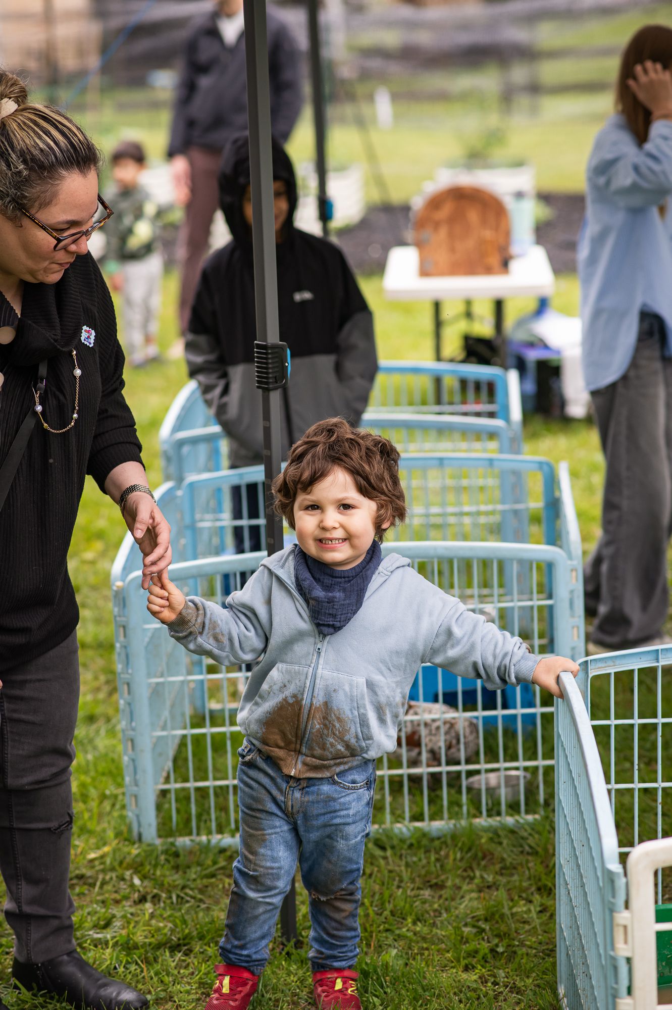Toddler with muddy clothes smiling and holding hands with a woman near small animal pens at an outdoor event.