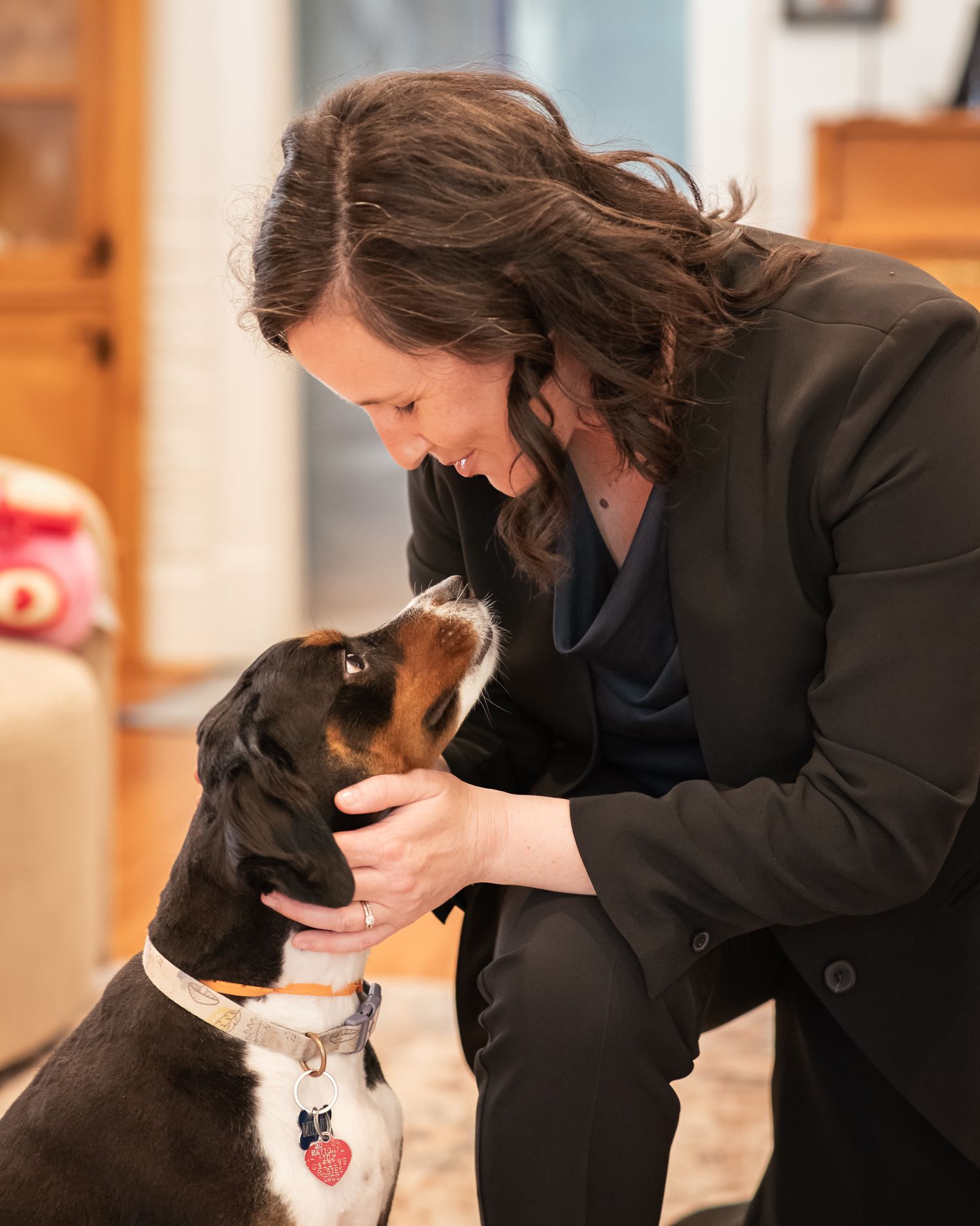 Woman in black blazer smiling and bonding with her brown and white dog indoors.