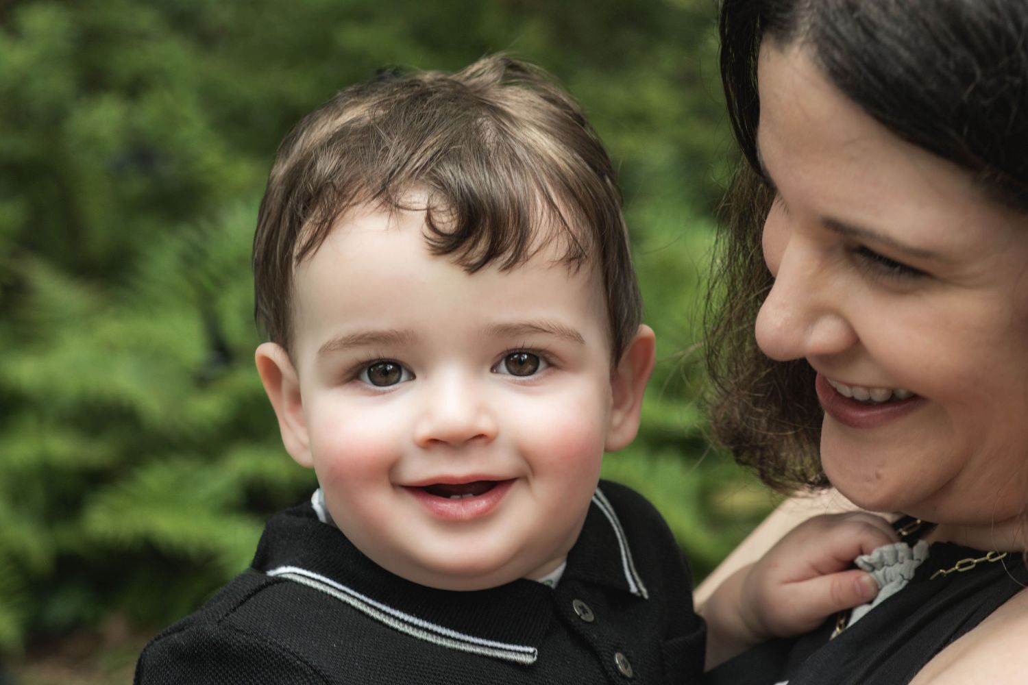 A smiling baby boy in a black polo shirt being held by a dark-haired woman outdoors near green trees.