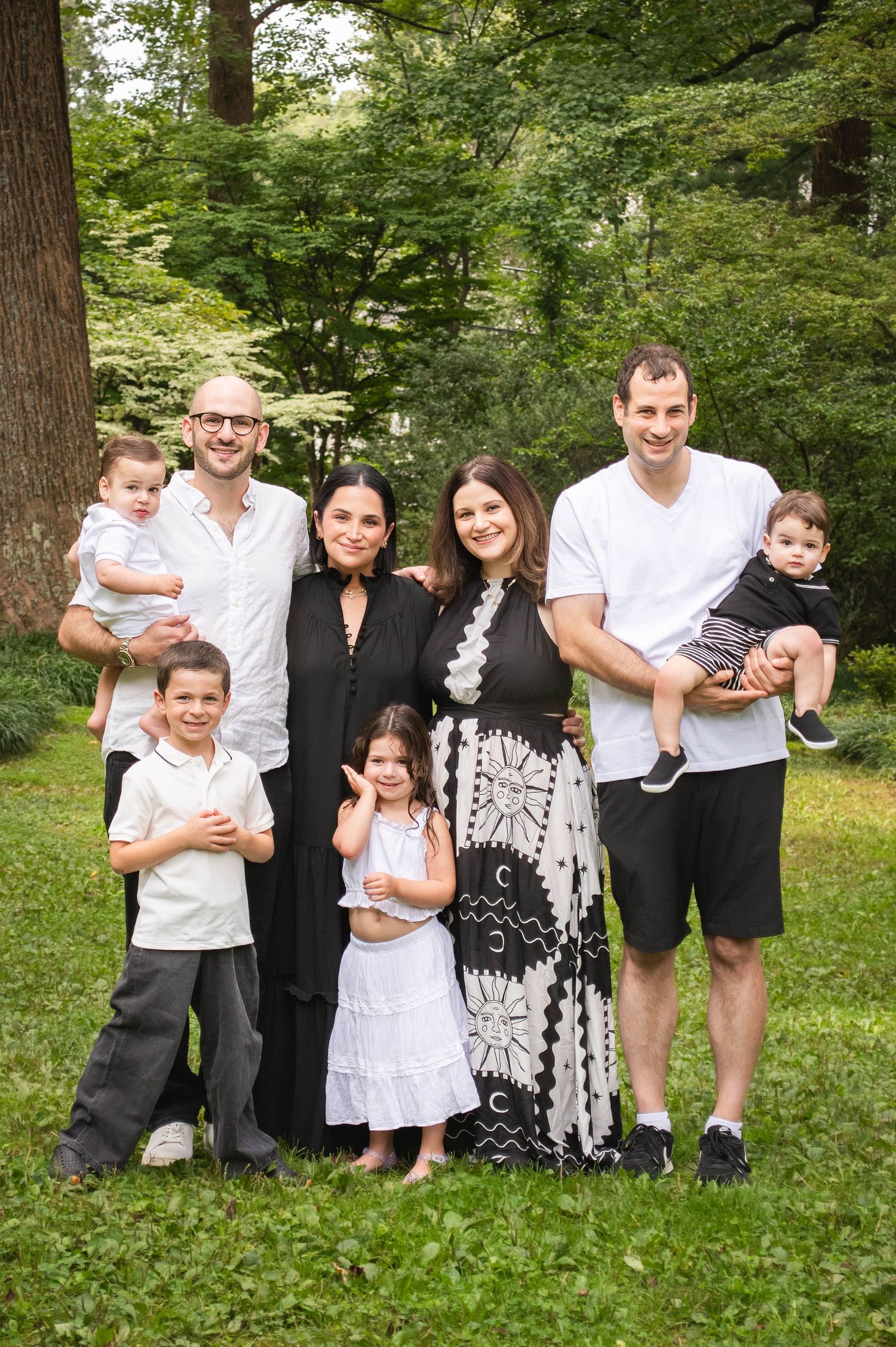 Family of seven posing outdoors in a lush green park, smiling together on a summer day.