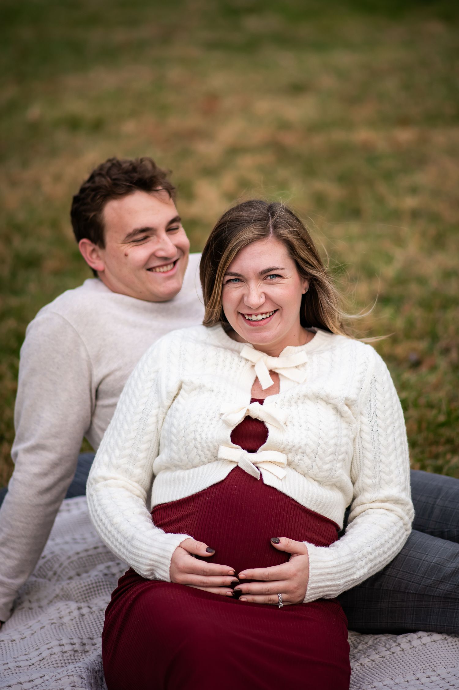 A couple in white sweaters and burgundy clothing lay together on a blanket outdoors sharing joyful moments.