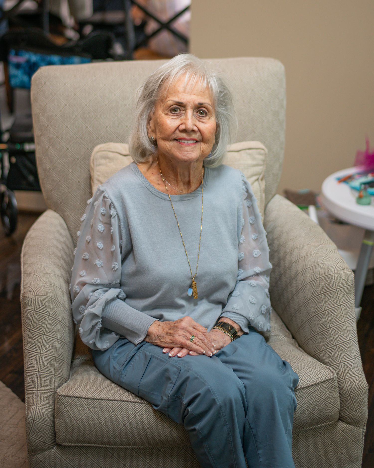 Elderly woman with white hair smiling in a beige armchair, wearing a blue outfit with a long necklace.