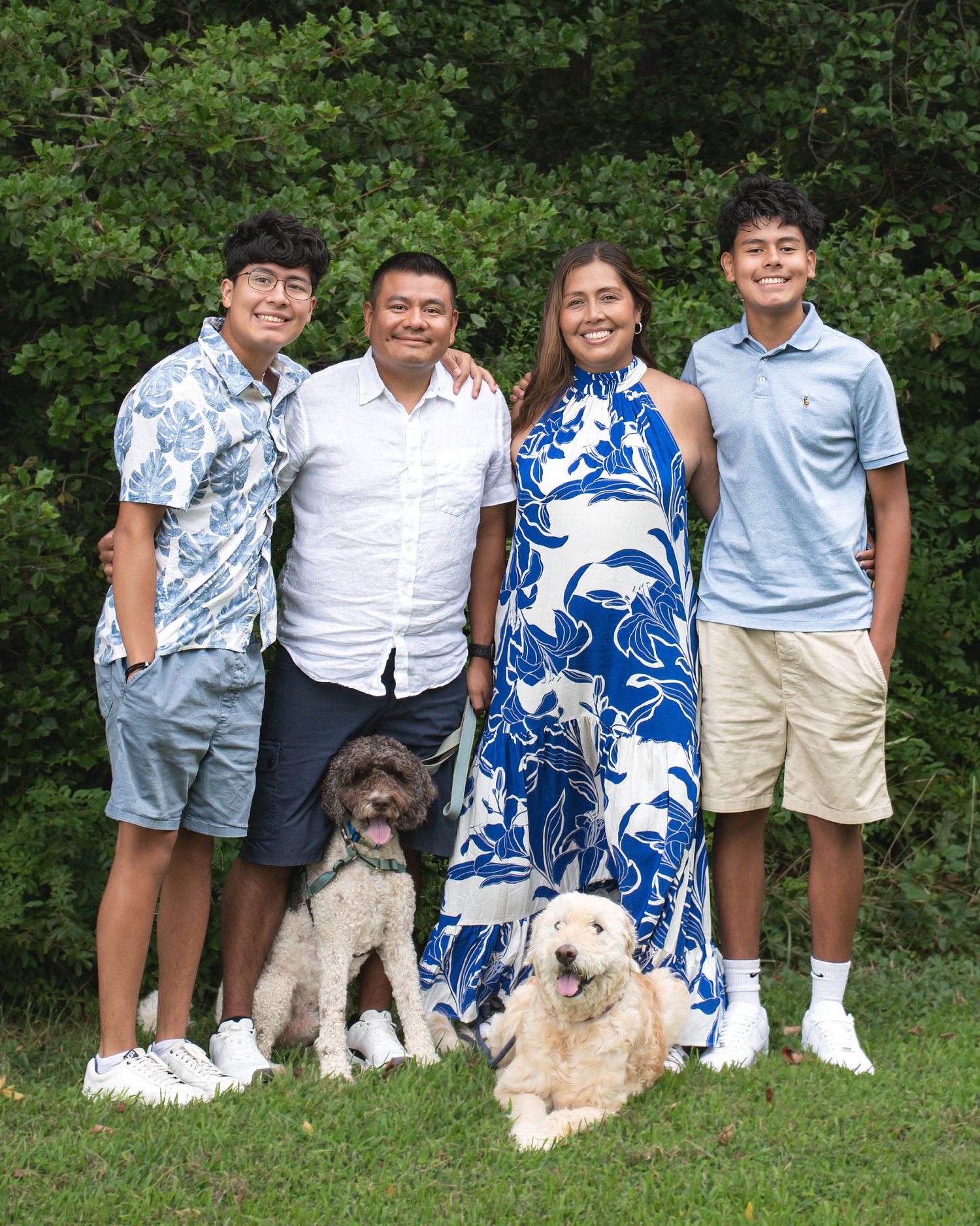 Family of four smiling outdoors with two fluffy dogs, surrounded by lush green trees, wearing coordinated casual outfits.