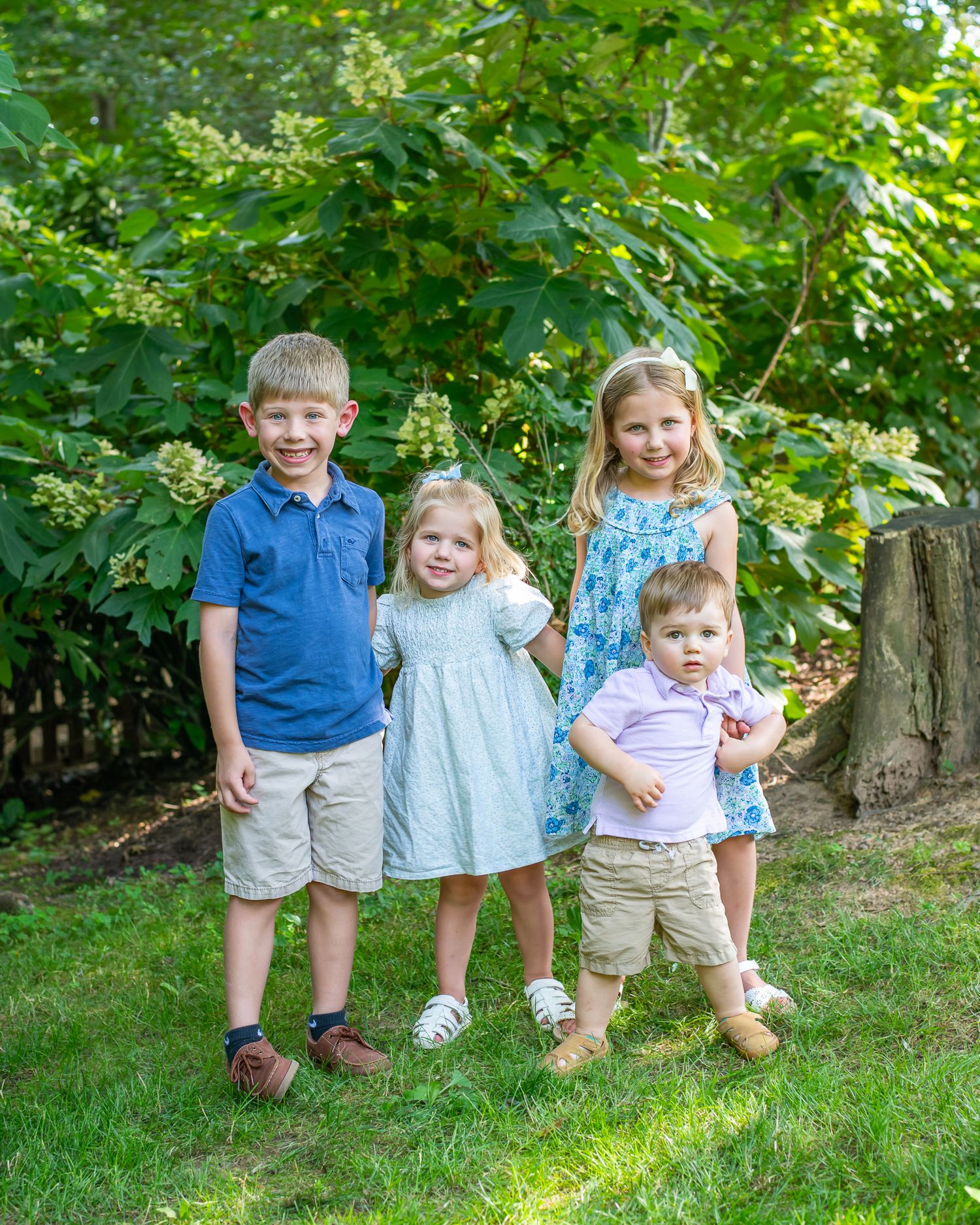 Four young children smiling together outdoors in a lush green garden, dressed in pastel summer outfits.