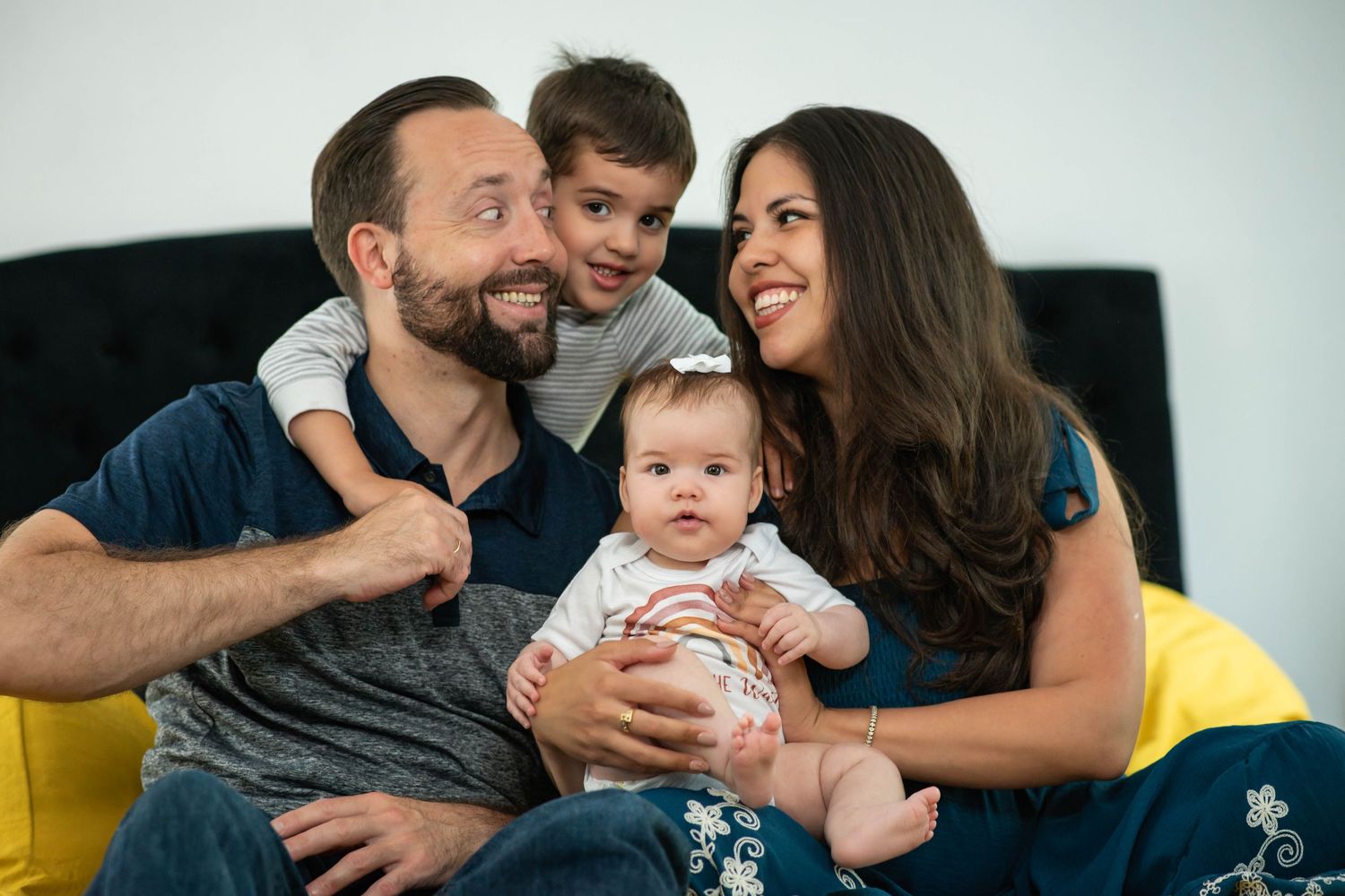 Happy family of four with dad, mom, toddler boy, and baby girl smiling together on a yellow chair.