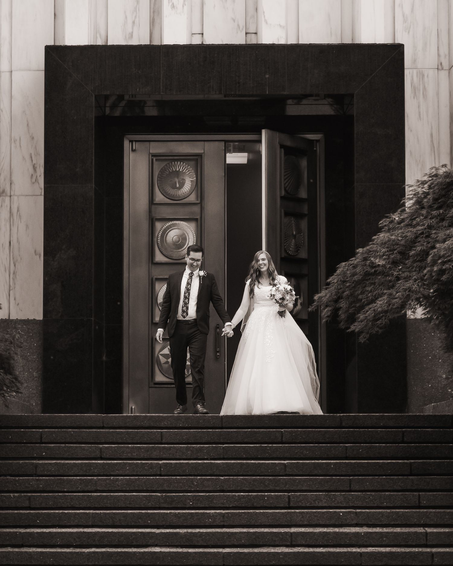 Black and white photo of a bride and groom holding hands on church steps after their wedding ceremony.