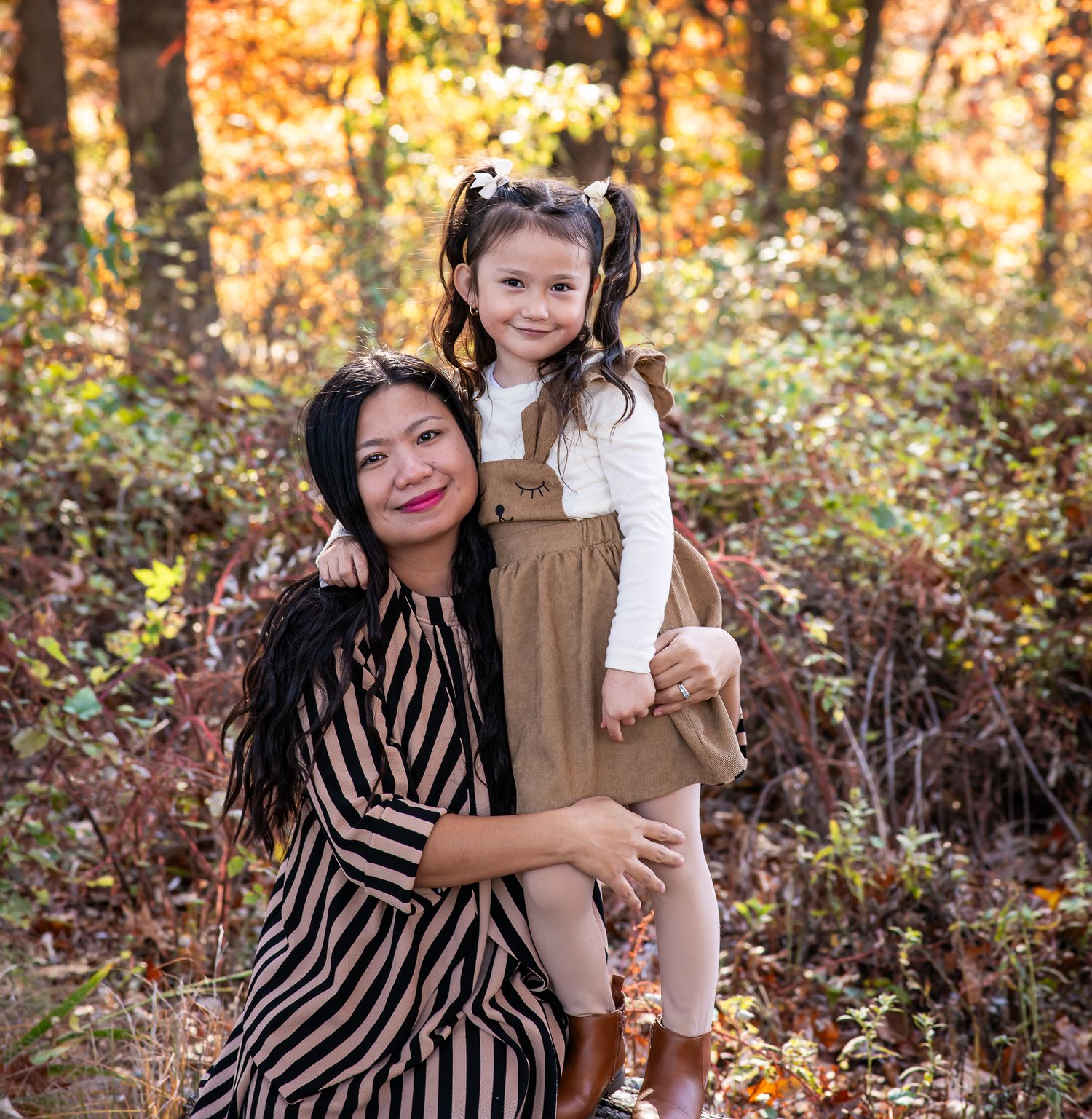 A mother holds her young daughter outdoors among fall foliage, both smiling in autumn attire.