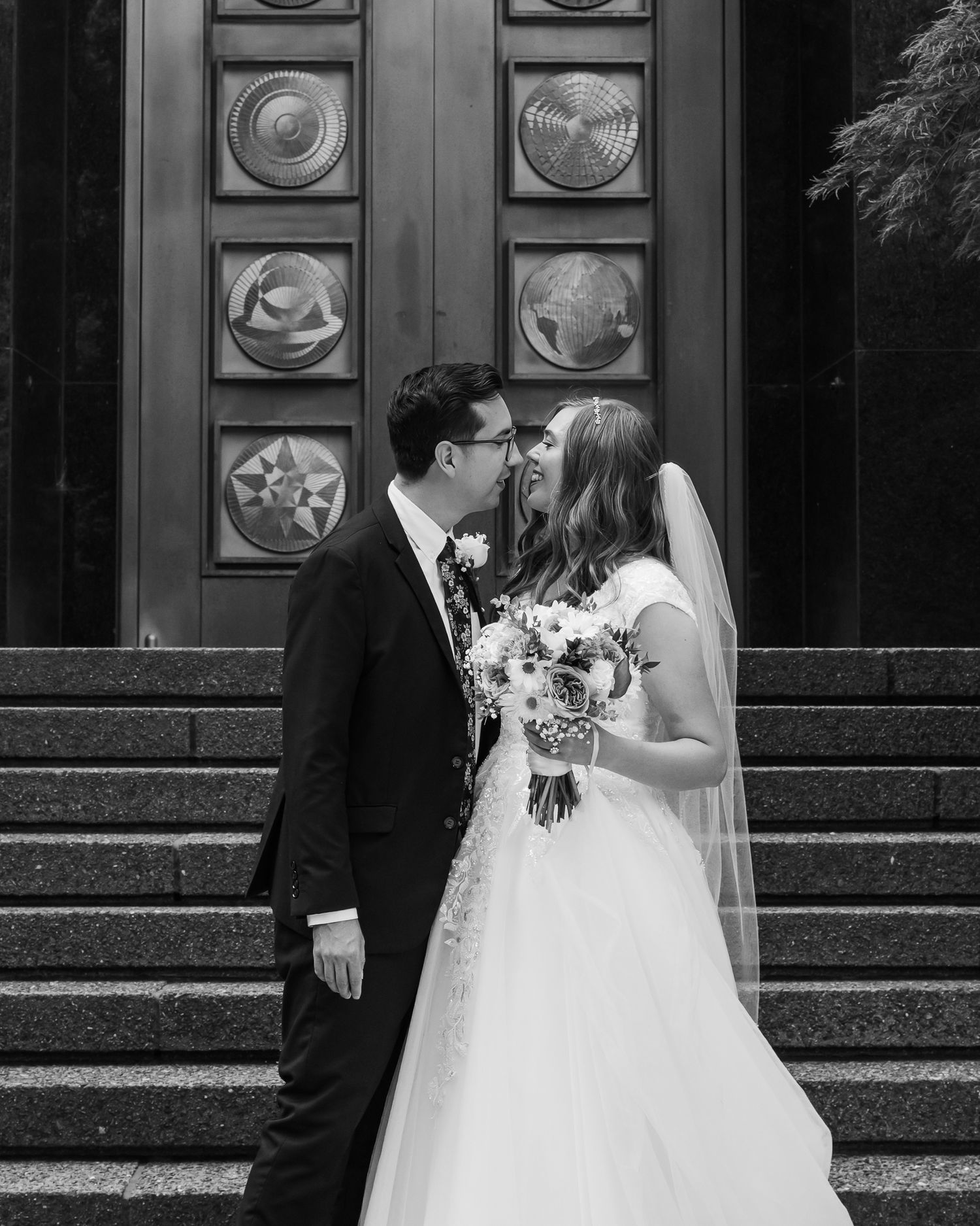 Black and white photo of a bride and groom sharing a kiss on church steps, bride holding a bouquet.