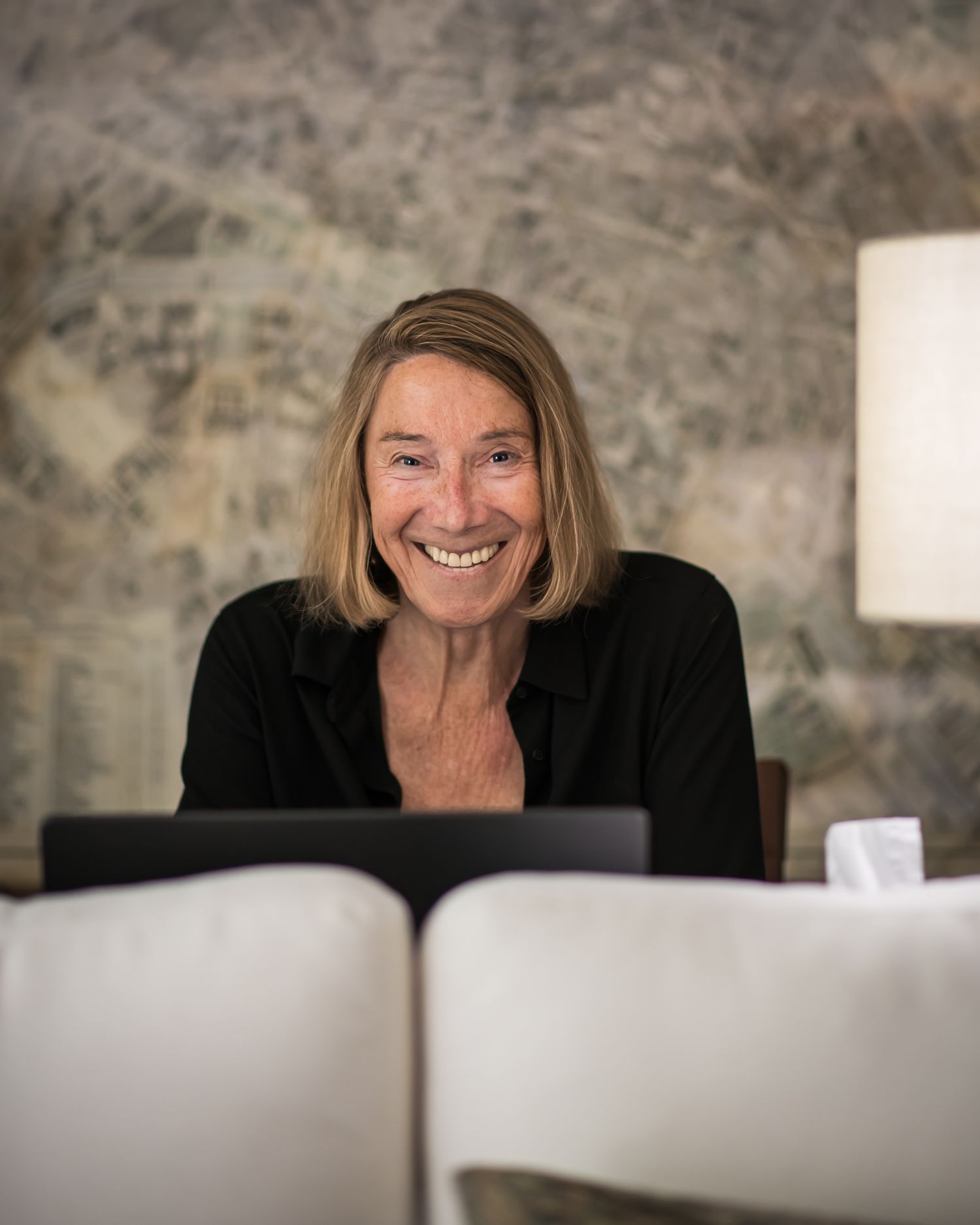 Smiling woman with blonde hair in black top seated indoors near stone wall and lamp.