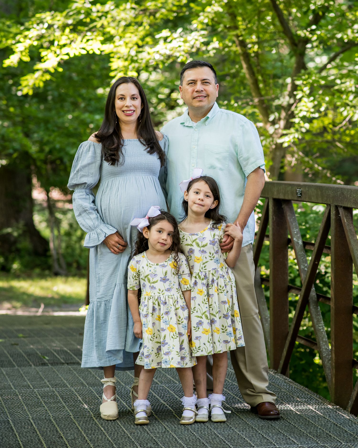 Pregnant woman in blue dress poses with husband and two young daughters in matching floral dresses on a garden bridge.