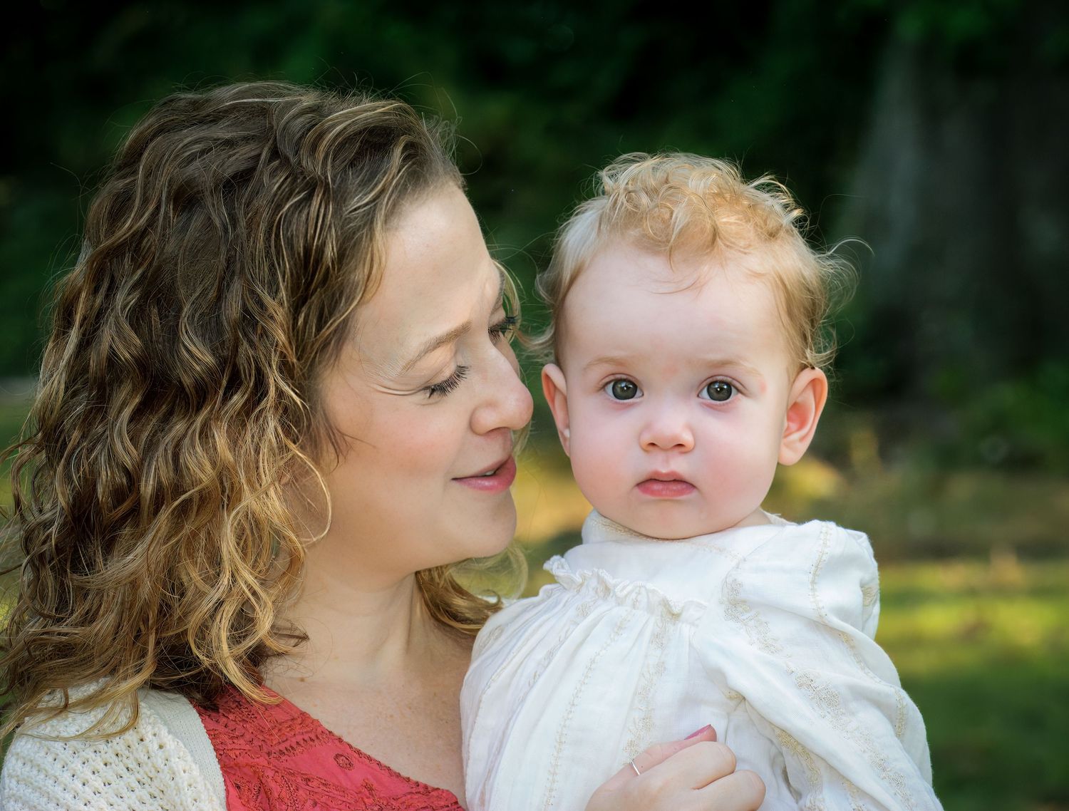 An intimate portrait of a parent holding a baby in white clothing against a green outdoor background.
