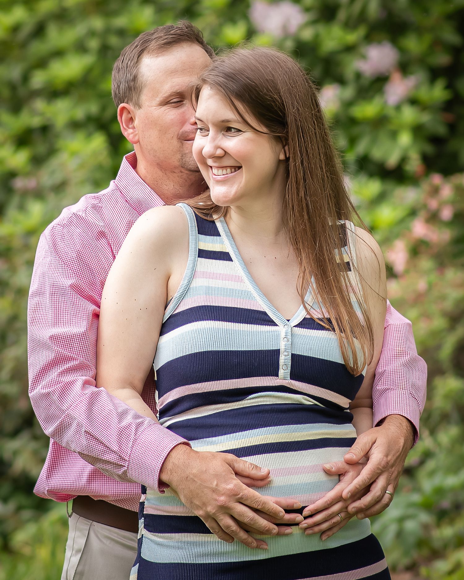 A couple shares a tender moment outdoors, with the man embracing a smiling pregnant woman in a striped top.