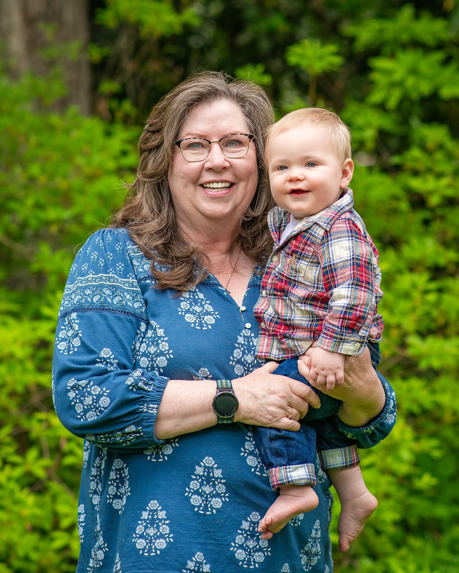 Smiling woman in blue floral dress holding a happy baby boy in plaid shirt outdoors.