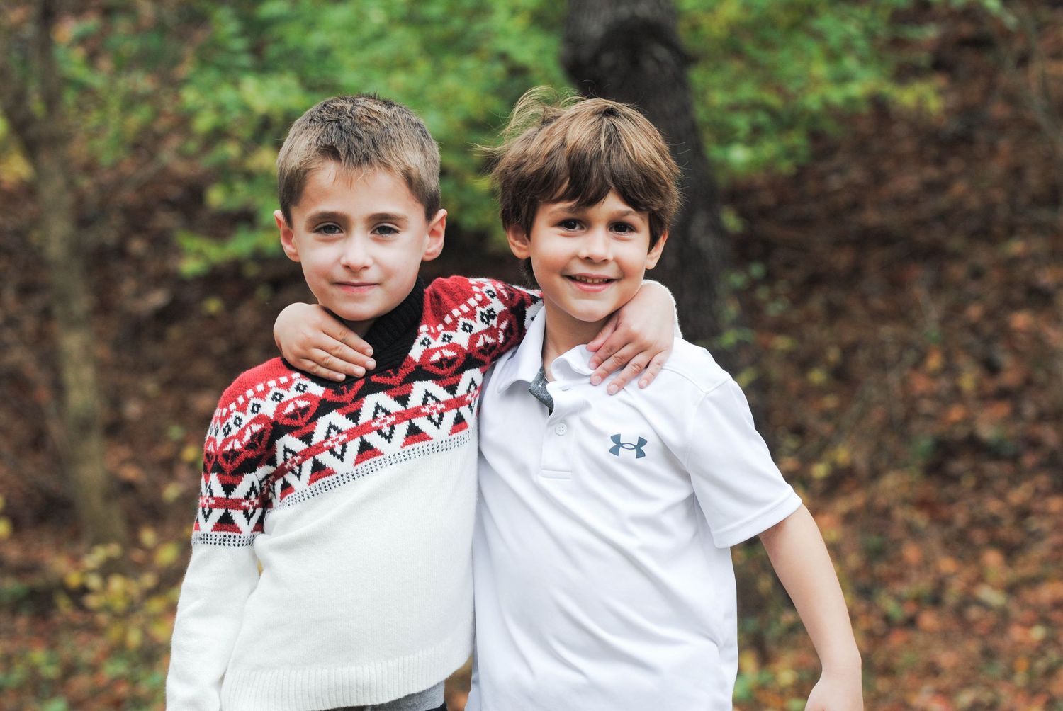Two young boys in winter sweaters smile together outdoors during fall.