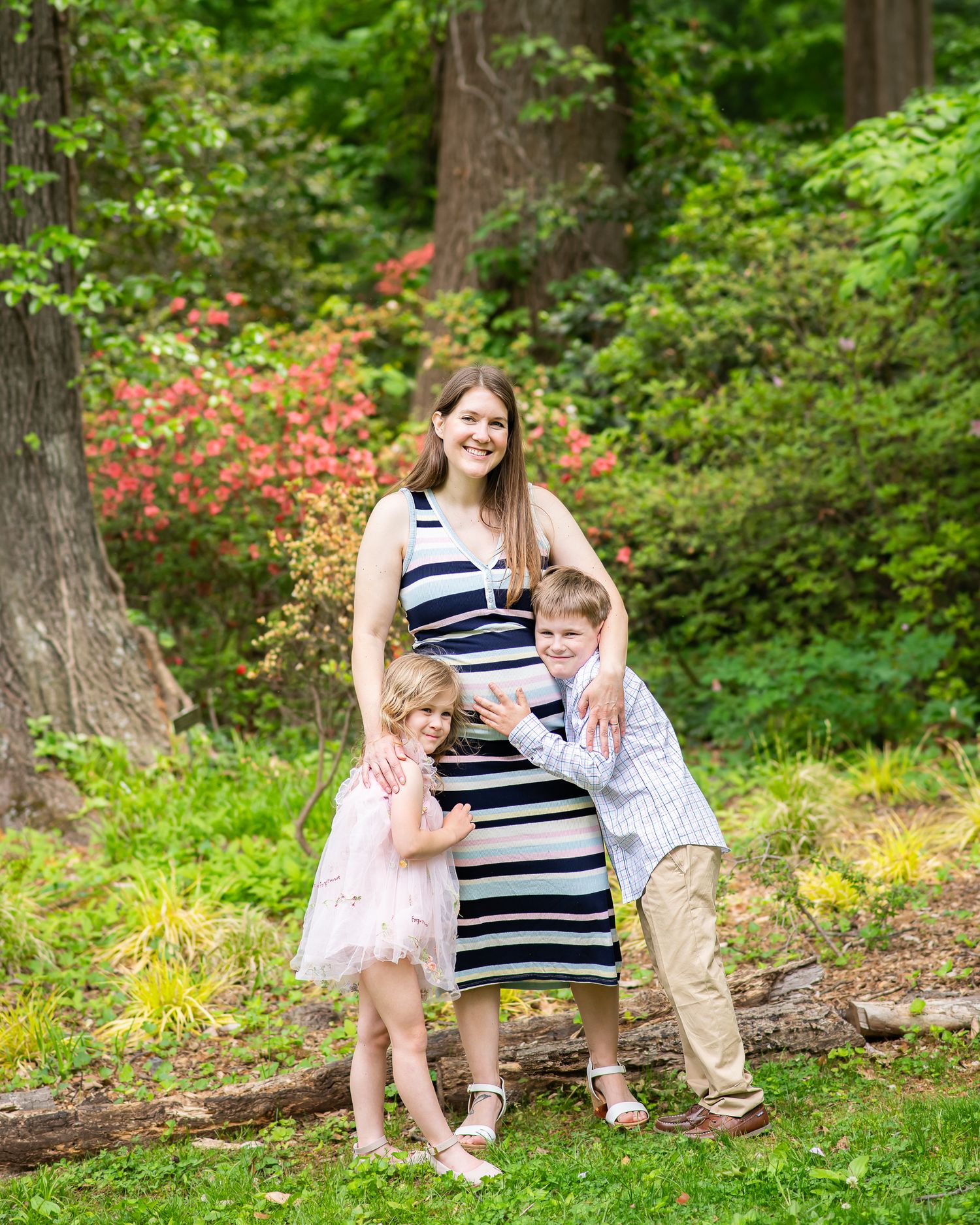 Pregnant woman in striped dress poses outdoors with two young children hugging her in a lush green garden setting.