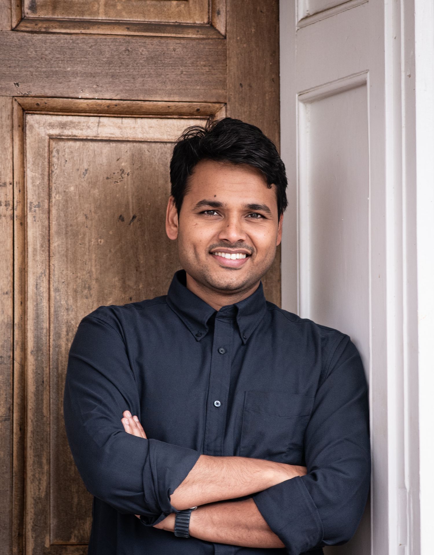 Professional portrait of someone in a navy button-down shirt standing confidently with arms crossed by a wooden door.