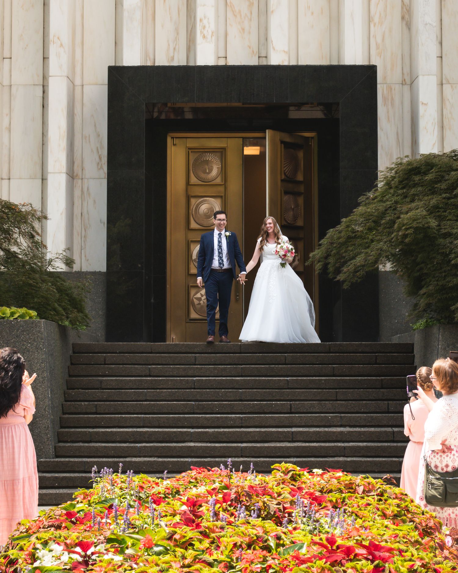 Bride and groom exit grand church doors, descending steps past colorful flower beds, as guests watch their joyful wedding ceremony.