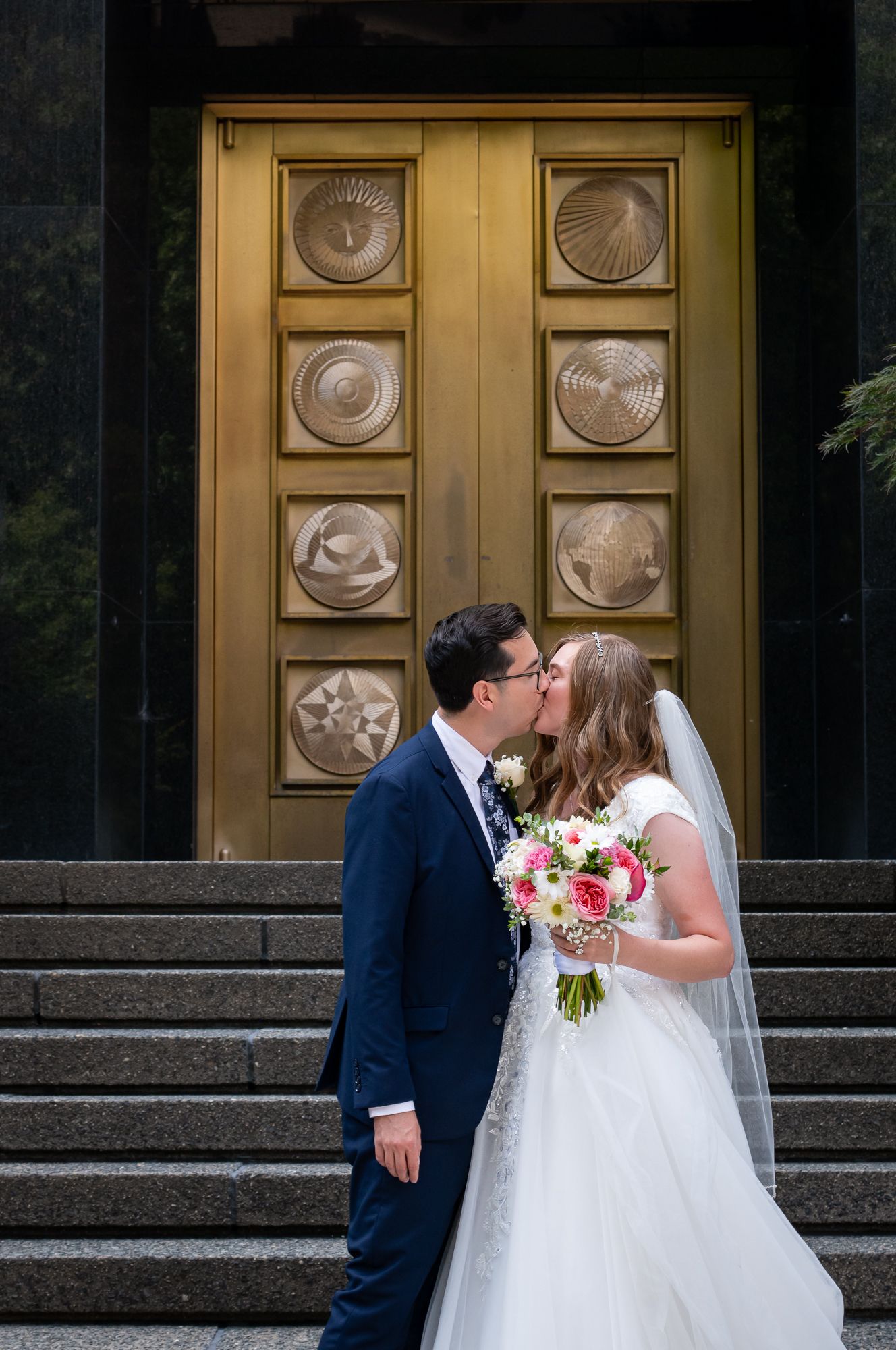 Bride and groom kissing on steps before ornate gold doors, bride holding pink and white bouquet wearing veil and white gown.