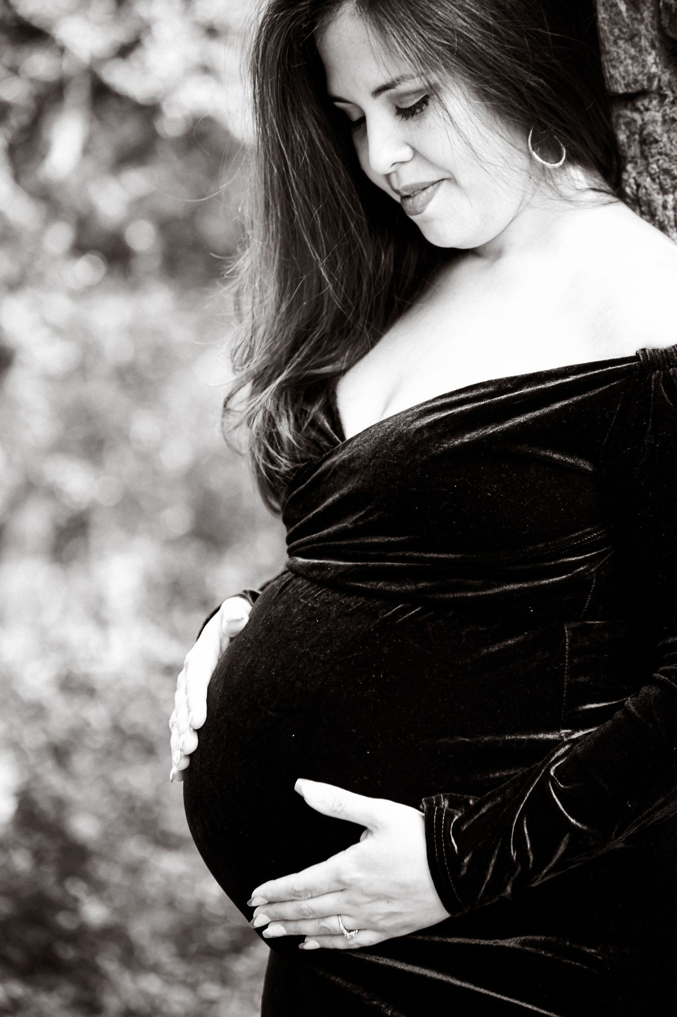 Black and white maternity photo of pregnant woman in velvet dress cradling baby bump outdoors.