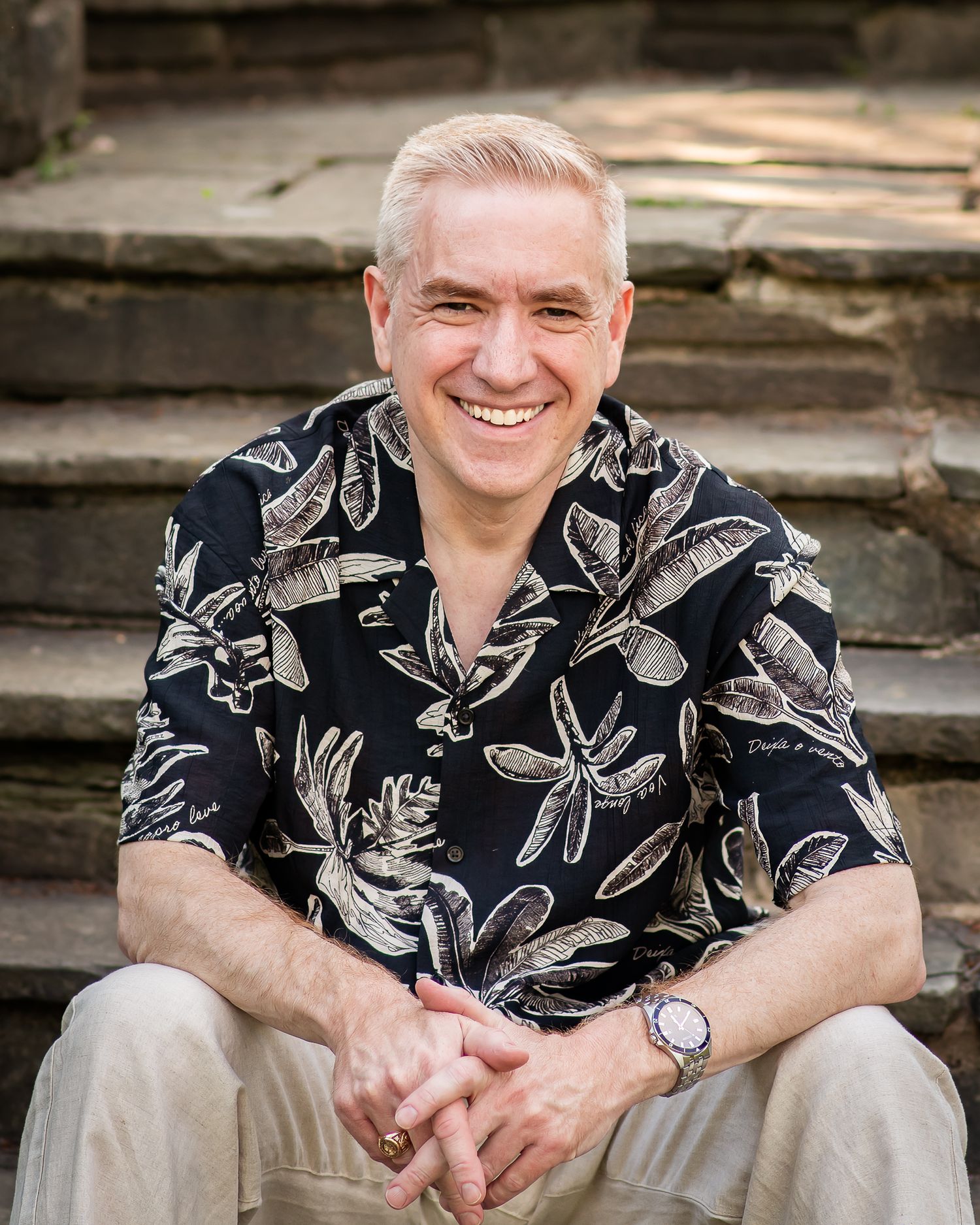 Smiling middle-aged man with blonde hair sitting on stone steps wearing a black floral Hawaiian shirt.