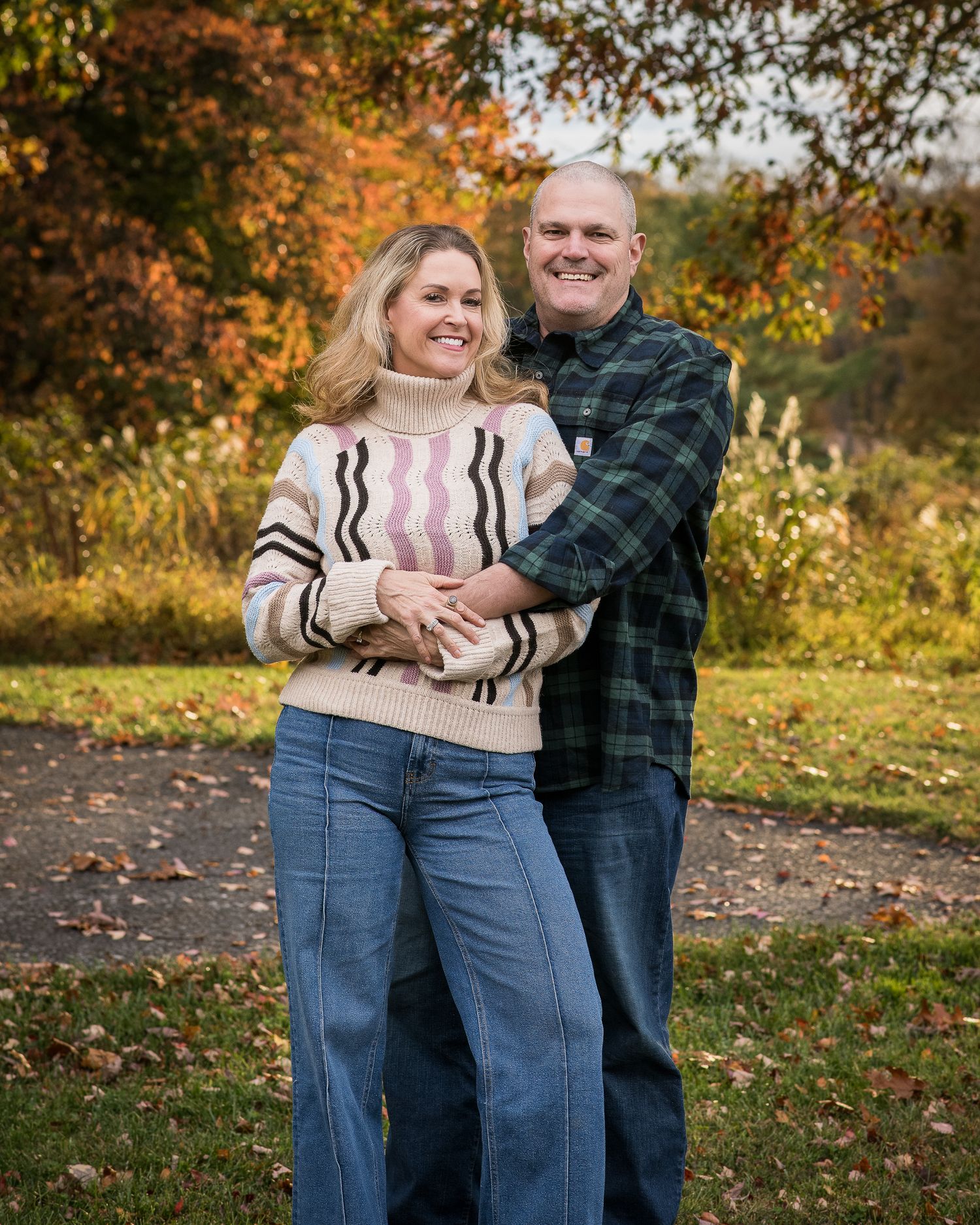 Smiling couple embraces outdoors in autumn park, woman in striped sweater and jeans, man in plaid shirt, fall foliage background.