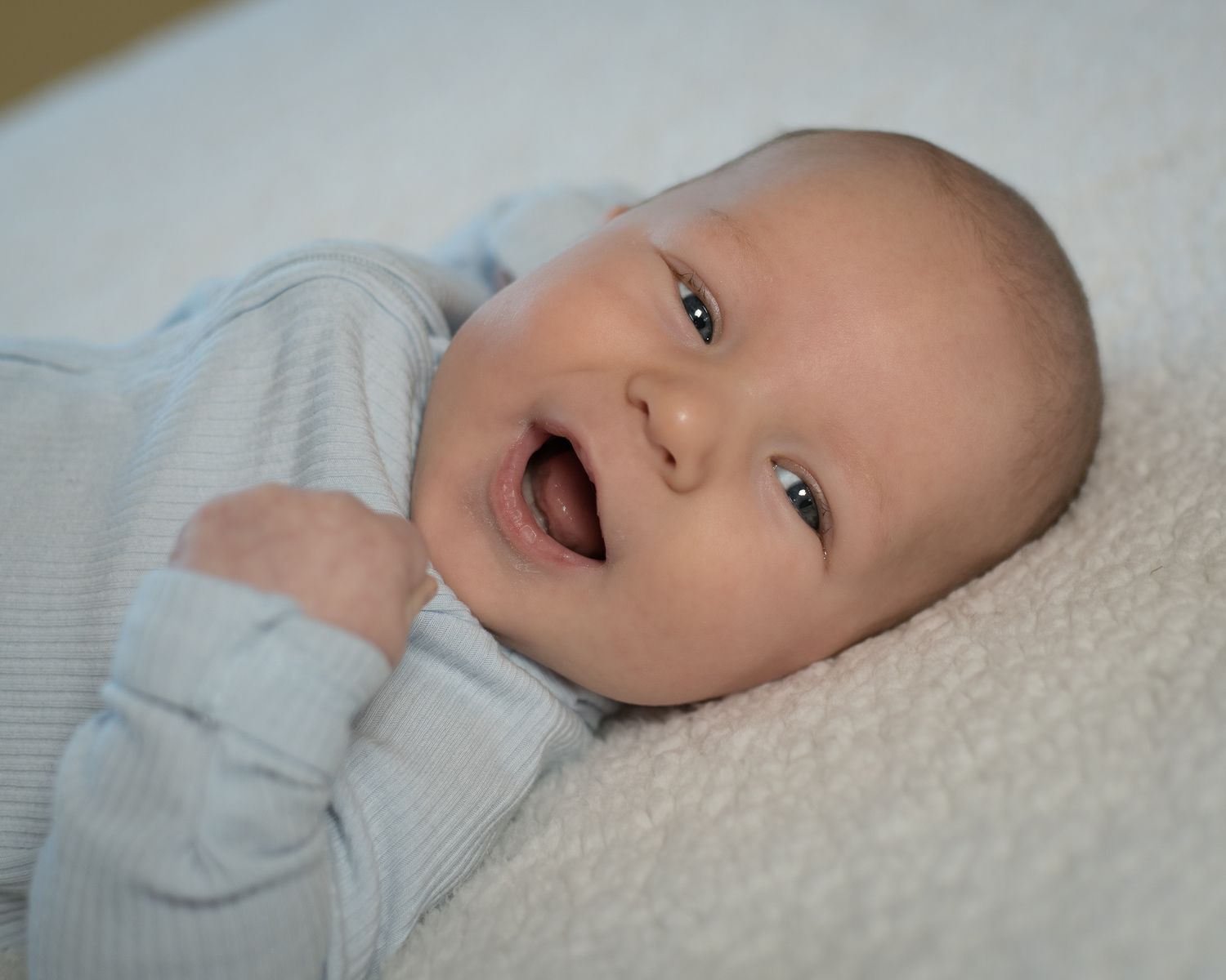 A smiling baby wearing a light blue outfit lies on a soft white blanket.