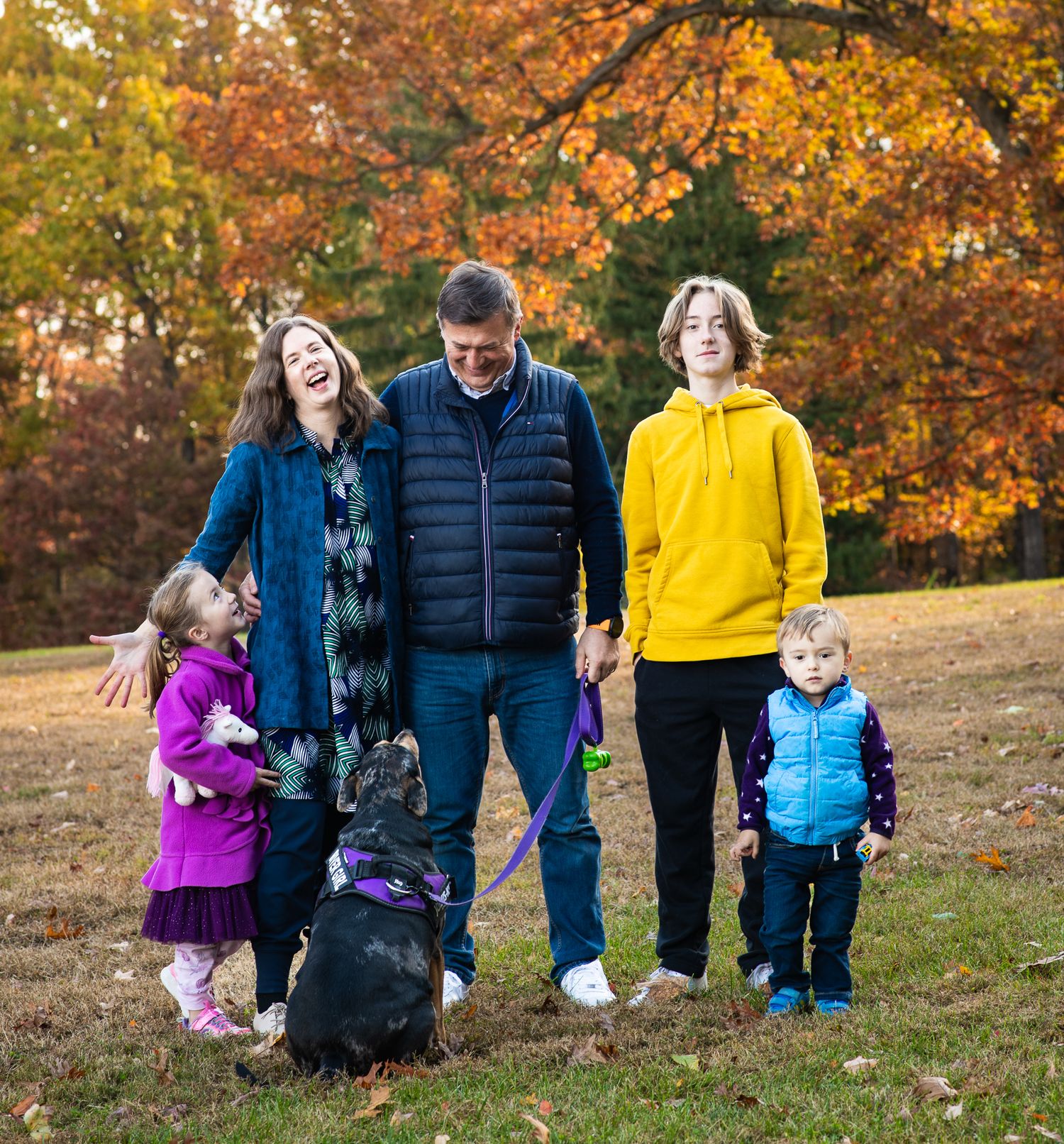 Family of five with dog enjoying autumn park, laughing and holding hands among colorful fall foliage.