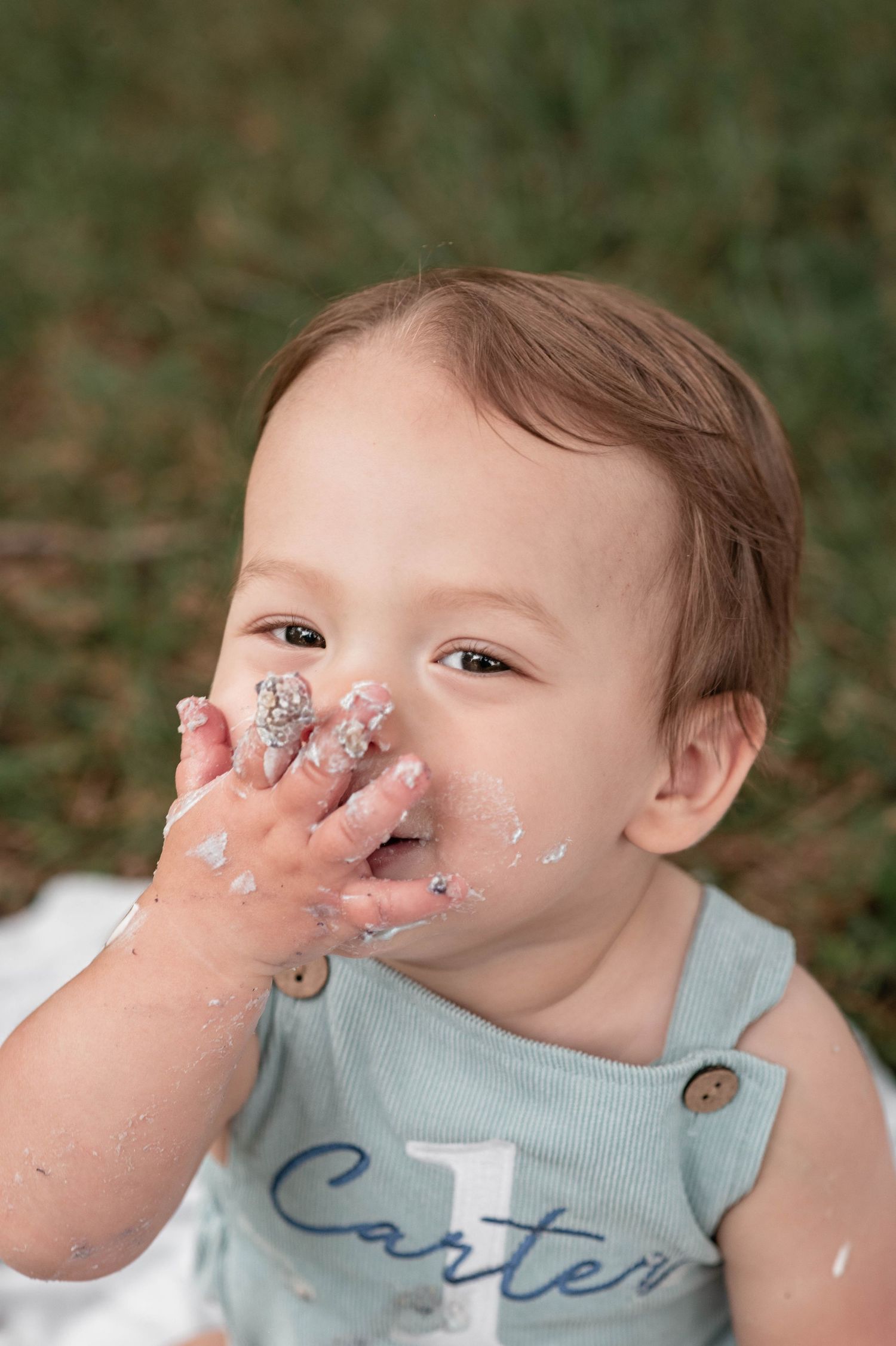 Baby Carter celebrates his first birthday with a cake smash, wearing a light blue romper outdoors.