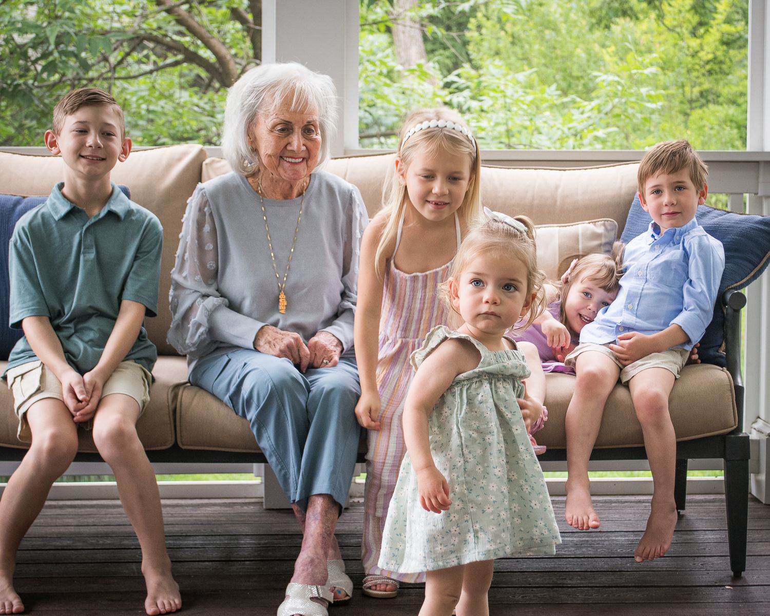 Elderly grandmother sits on patio sofa surrounded by five young grandchildren, smiling together in a warm family portrait.