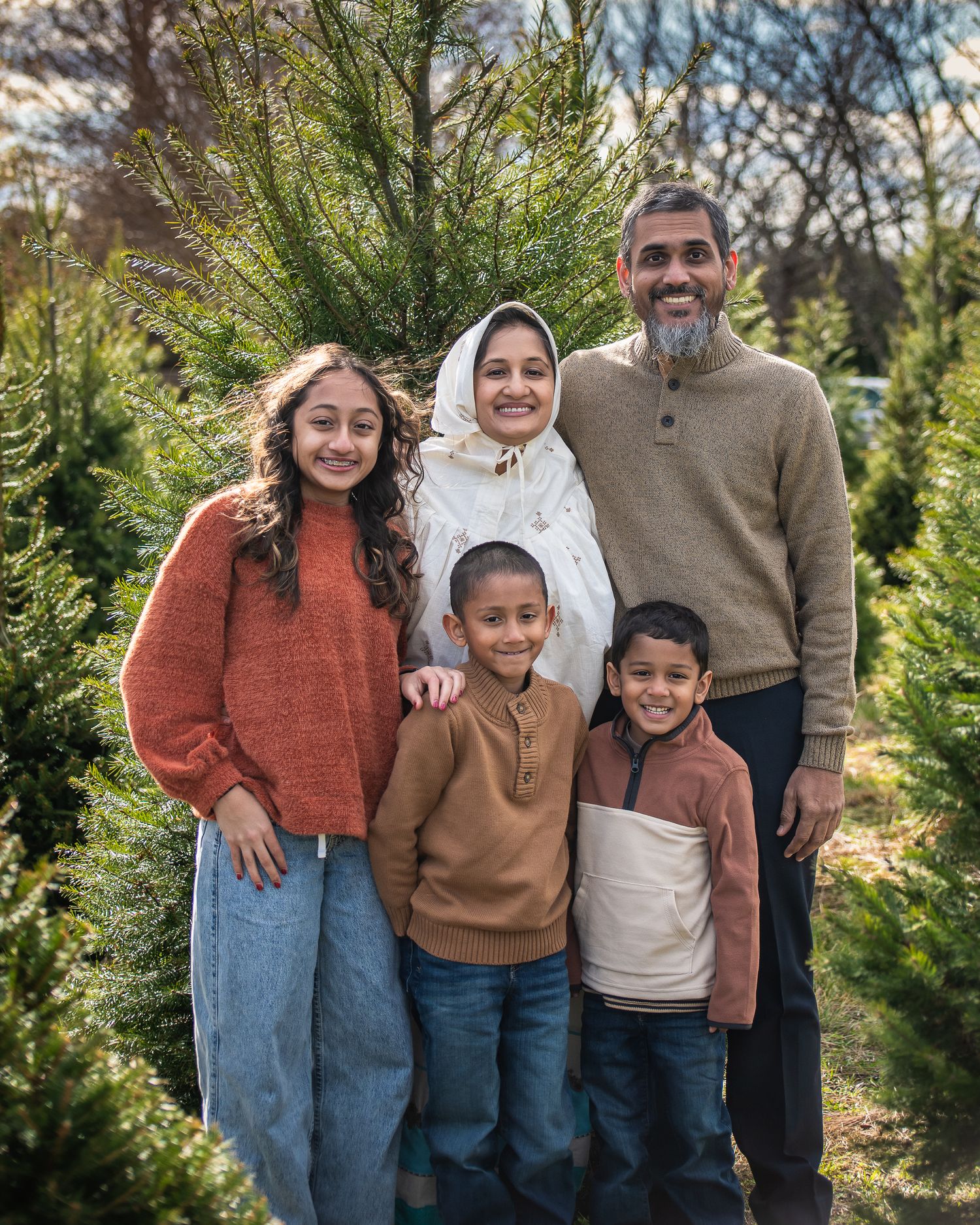 A family poses together outdoors among evergreen trees at a Christmas tree farm on a sunny day.