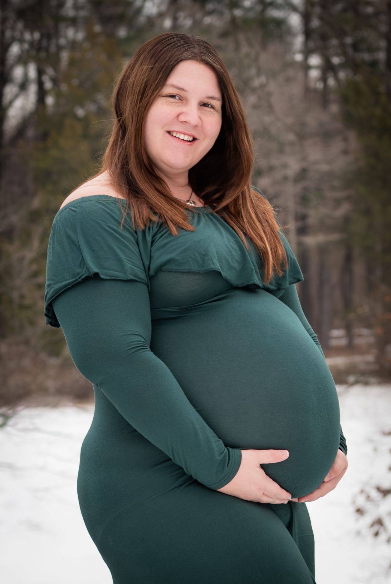 A person in a green long-sleeved dress poses for a maternity photo in the snow with trees in the background.