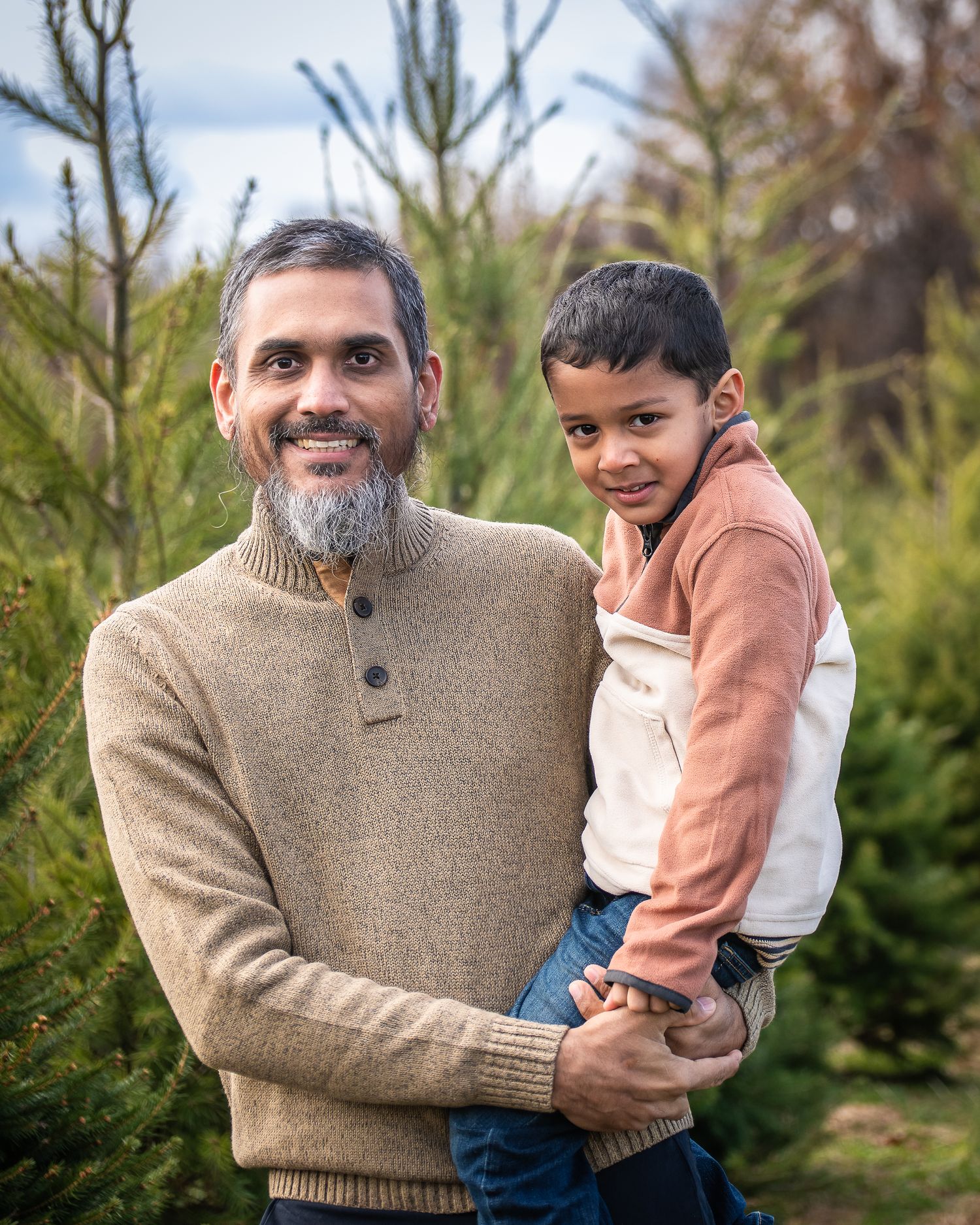 A parent and child share a joyful moment together while standing among evergreen trees in an outdoor setting.