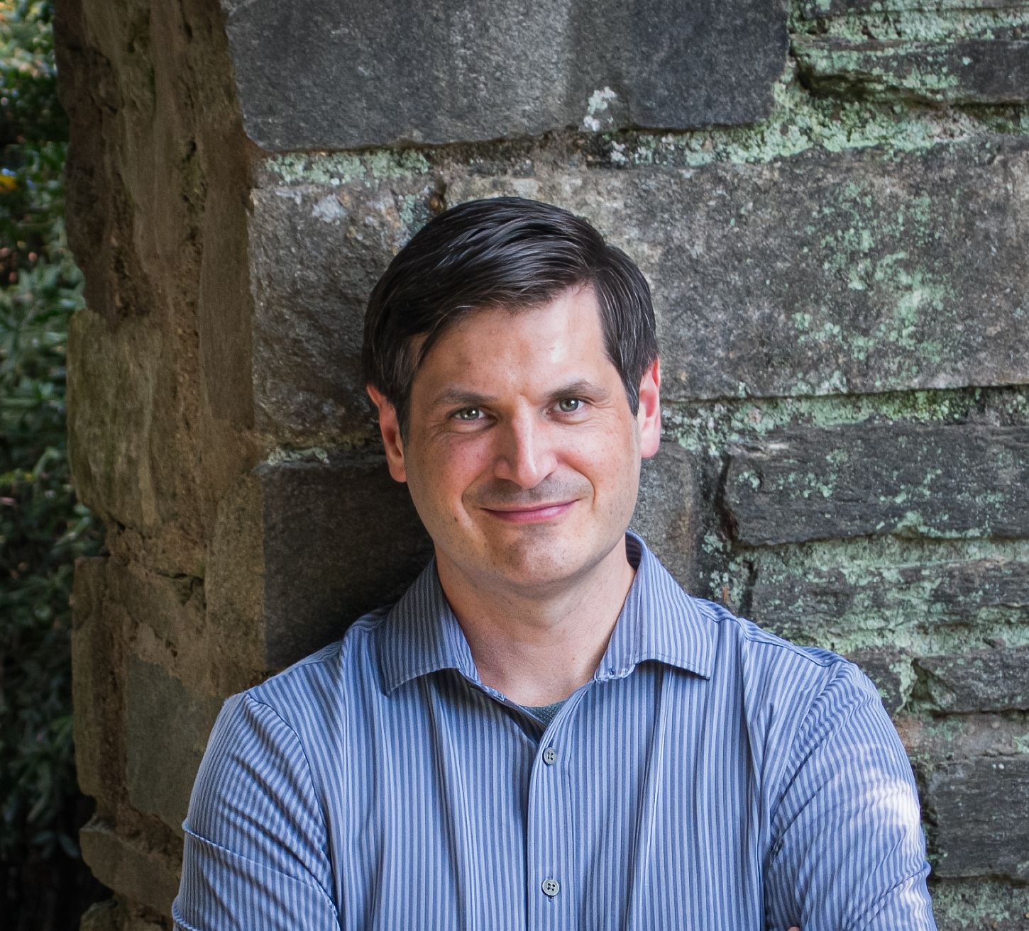 Professional headshot of someone in a blue striped button-down shirt against a stone wall background.